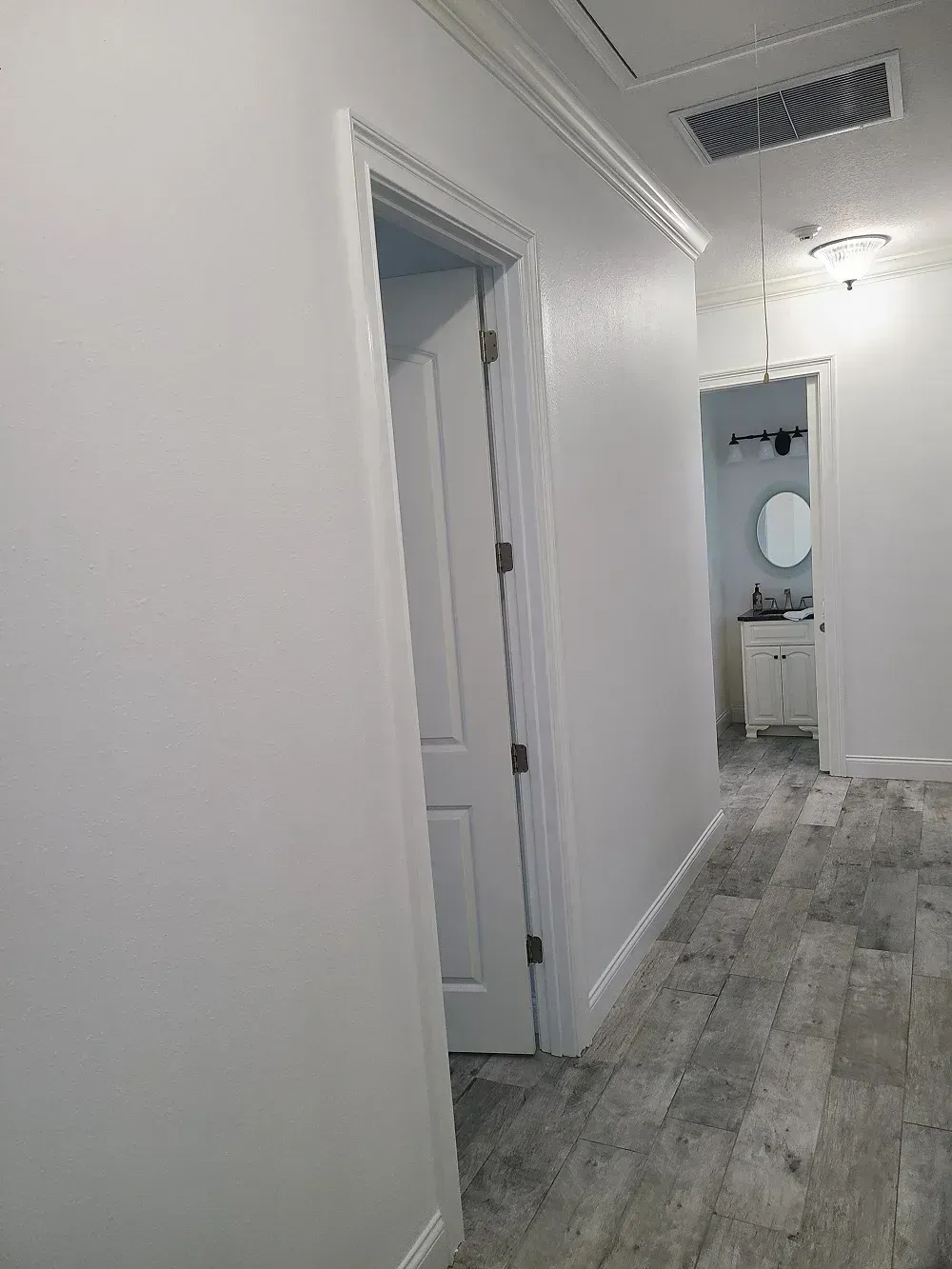 Empty hallway with white walls, gray wood floors, and open doors leading to a bathroom.