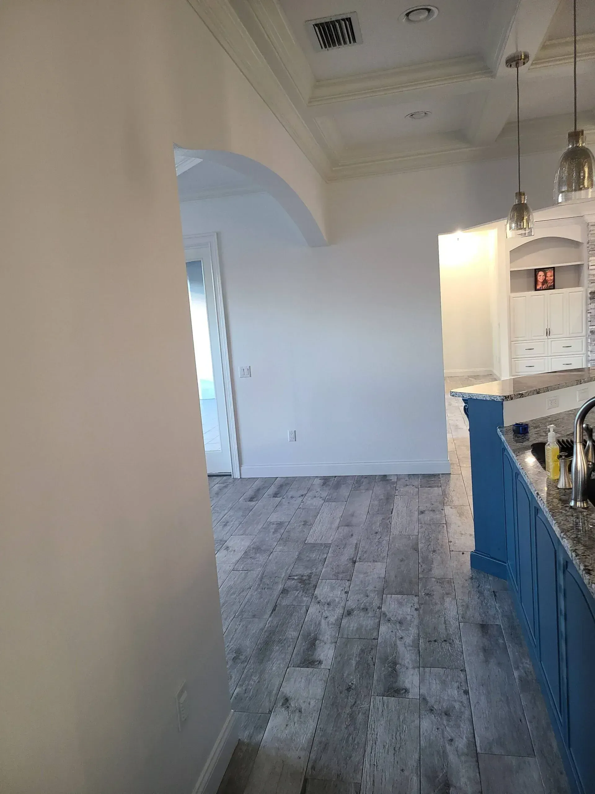 Modern kitchen hallway with gray wood floors, blue cabinets, and white walls leading to an open room