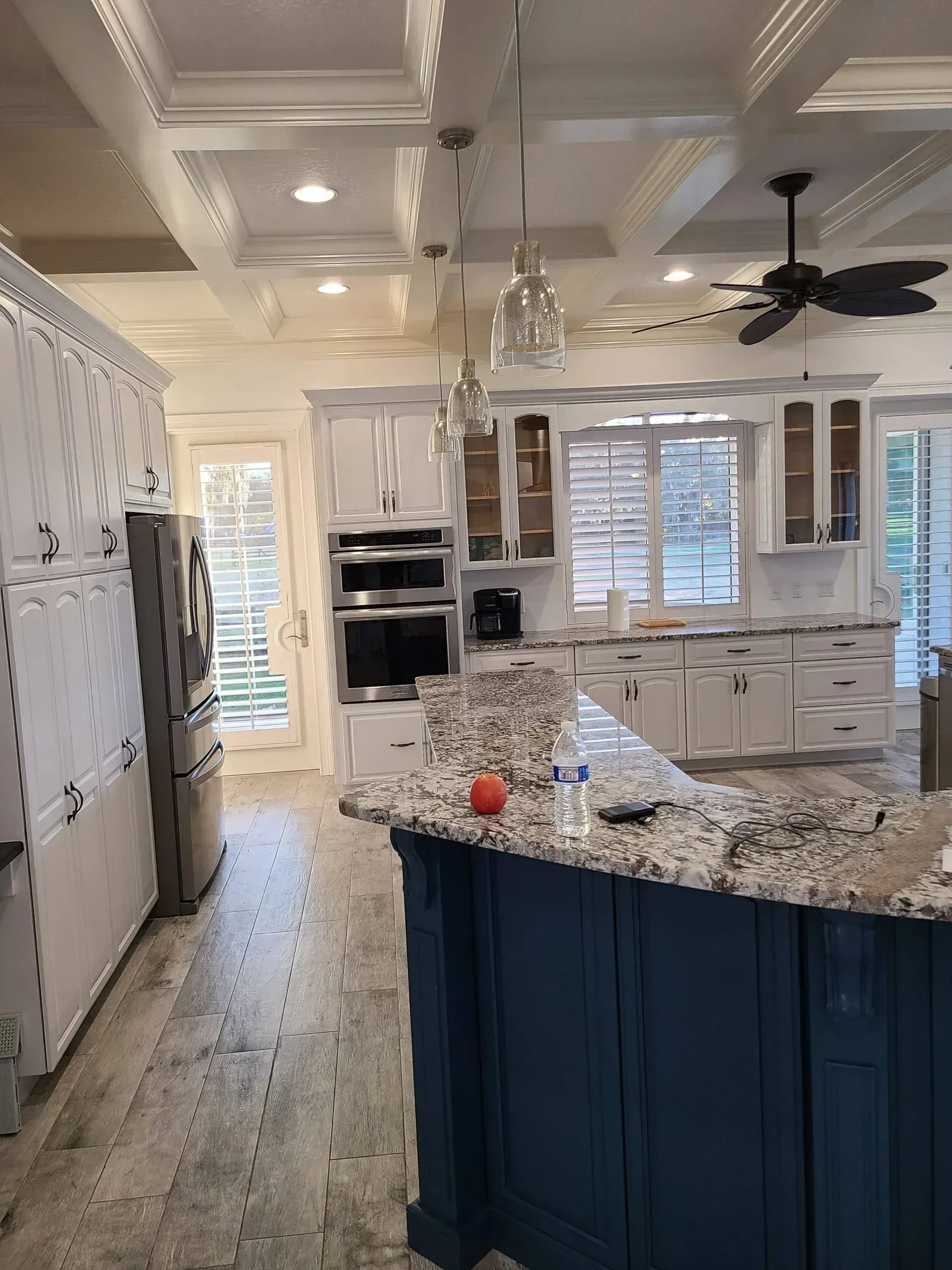 Bright kitchen with granite island, blue cabinets, white ceiling beams, and large windows.