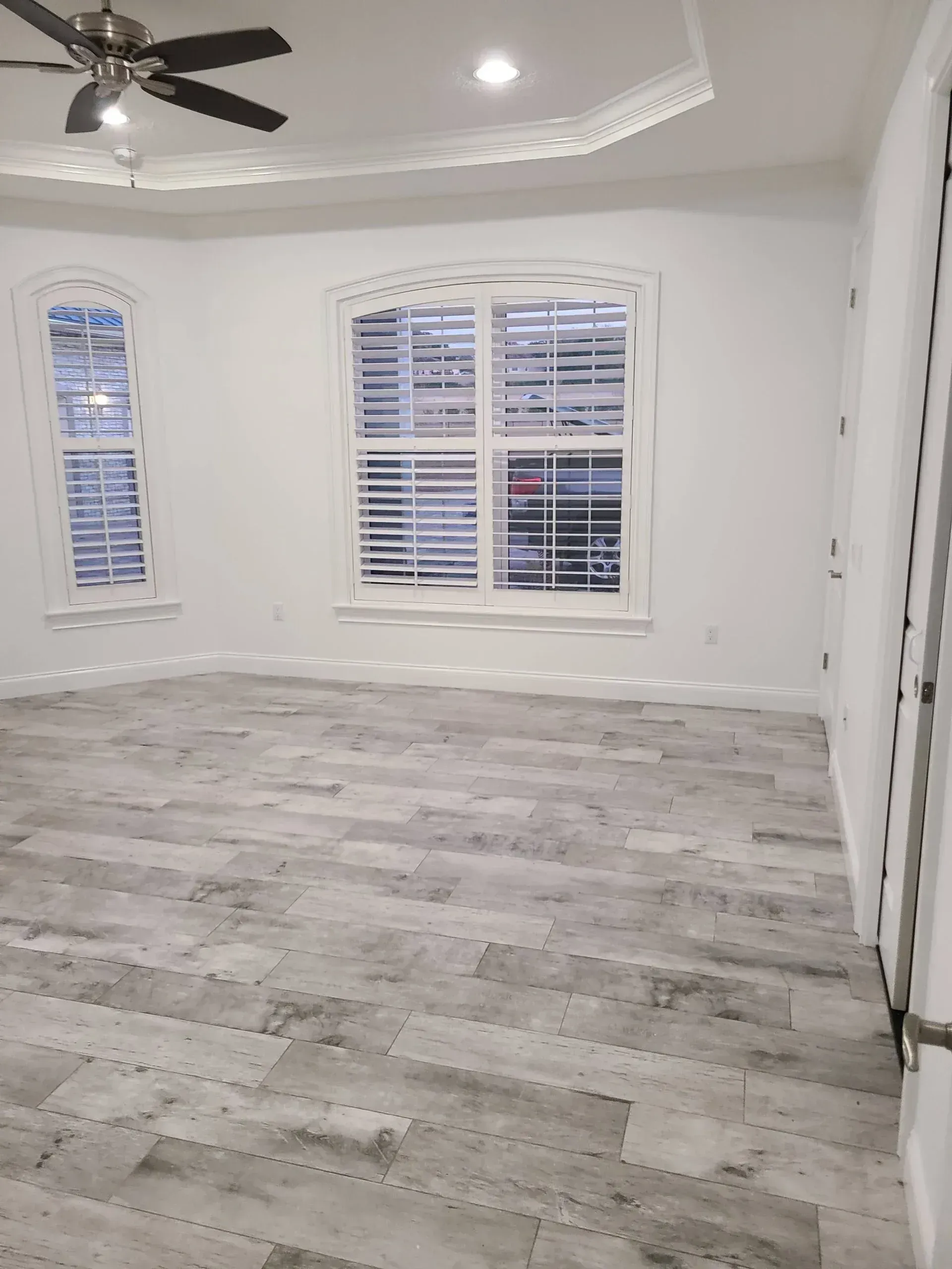 Empty bright room with gray wood-look floors, white walls, windows, and a ceiling fan.