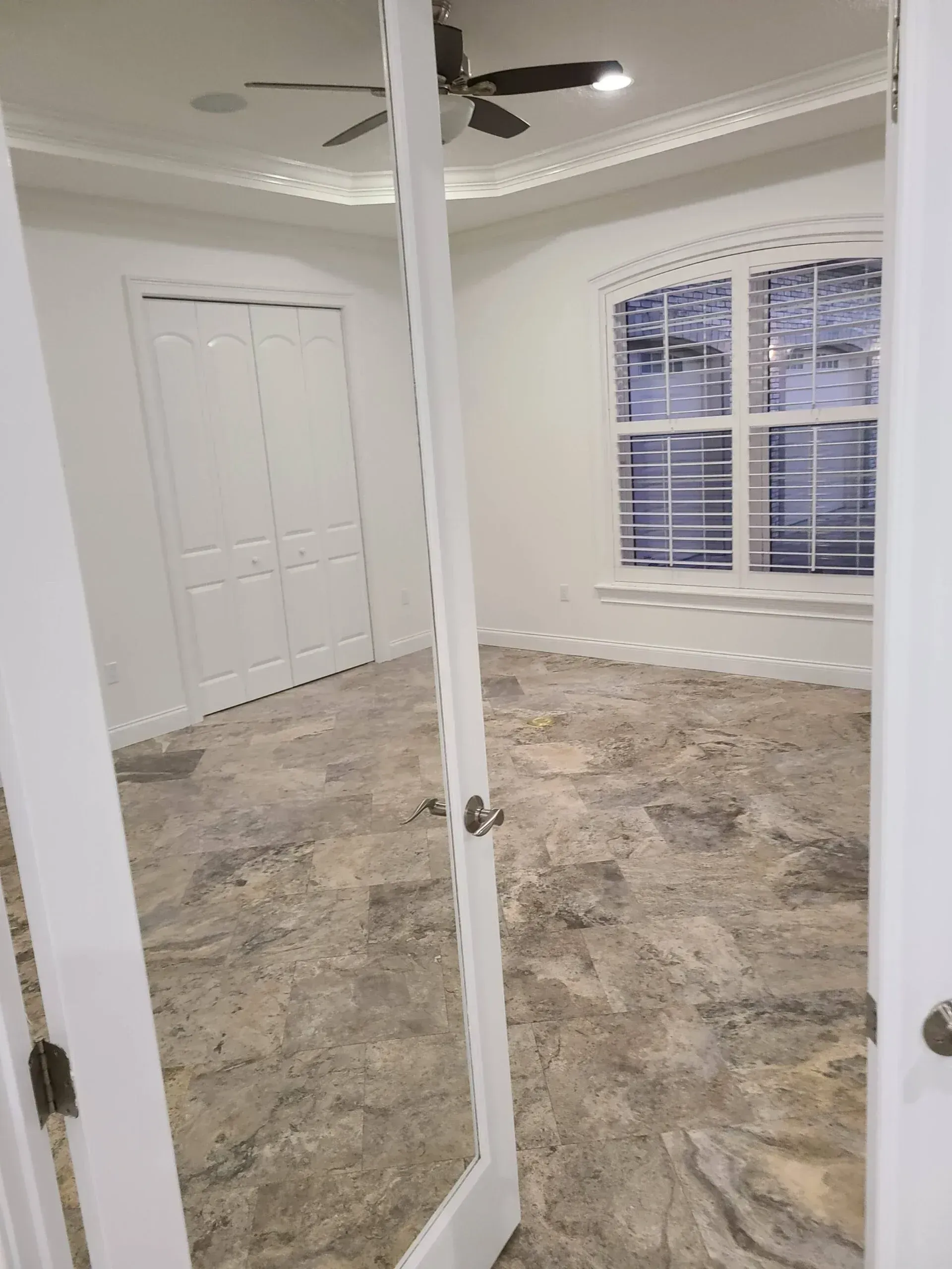 Empty bedroom with beige marble-pattern tile, white French doors, and a ceiling fan.
