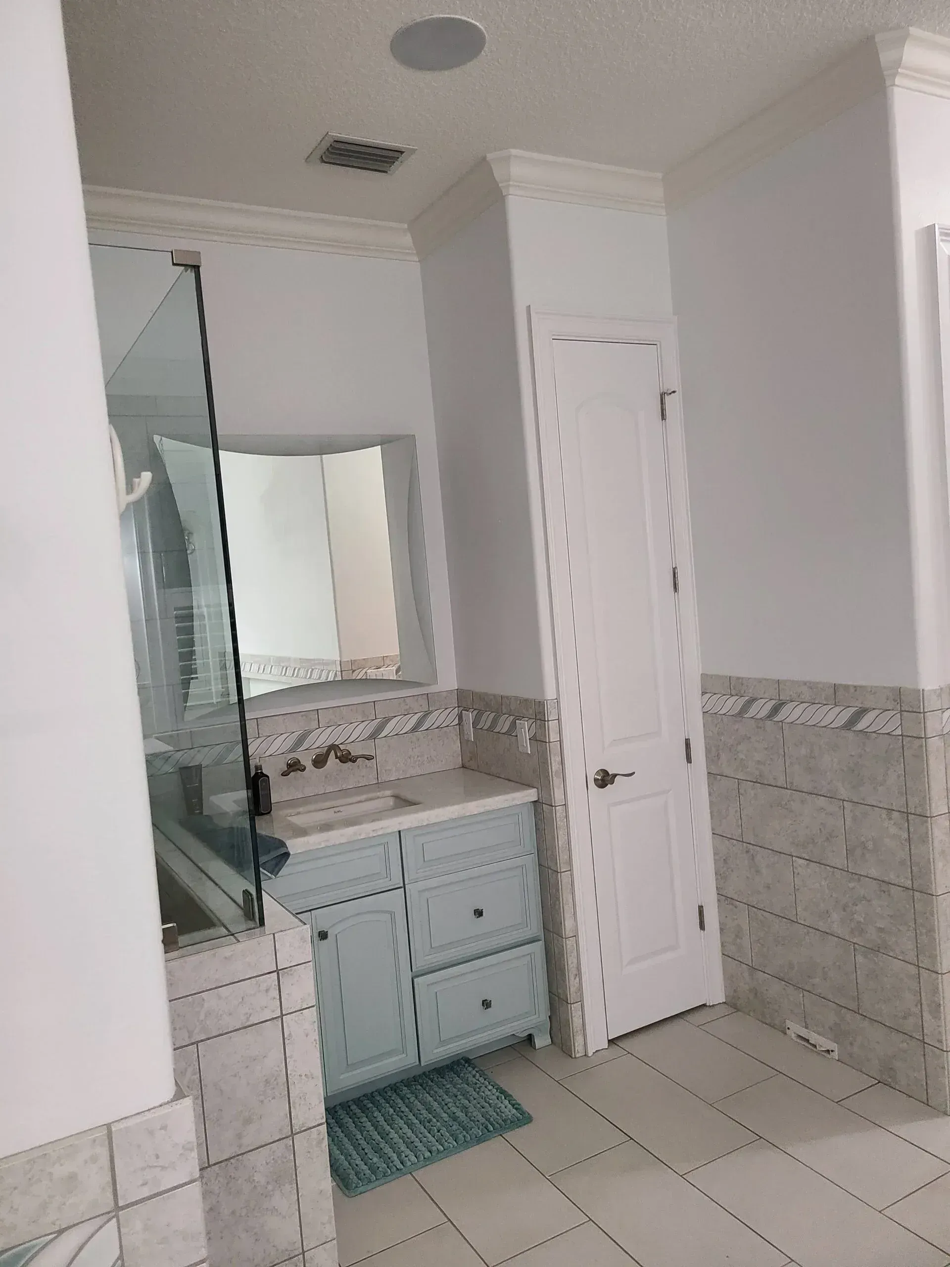 Bathroom with white tile floor, vanity, mirror, glass shower door, and a small teal rug.