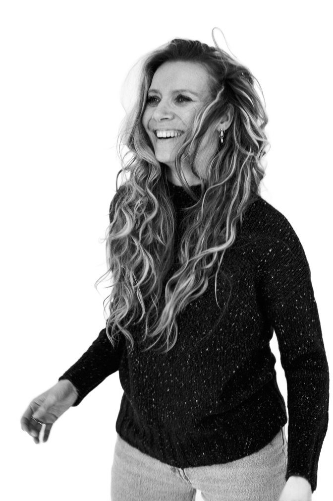 Debbie glimlacht en kijkt opzij, gekleed in een donkere trui, tegen een witte achtergrond.