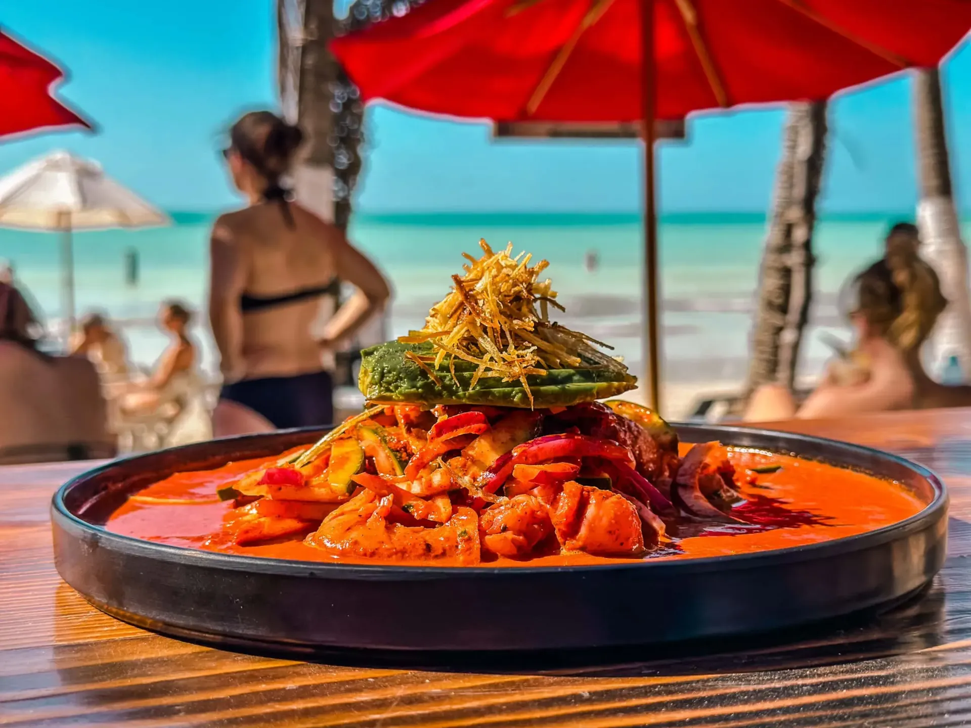 Plato de mariscos en salsa roja, cubierto con aguacate verde y guarnición crujiente, playa de fondo.