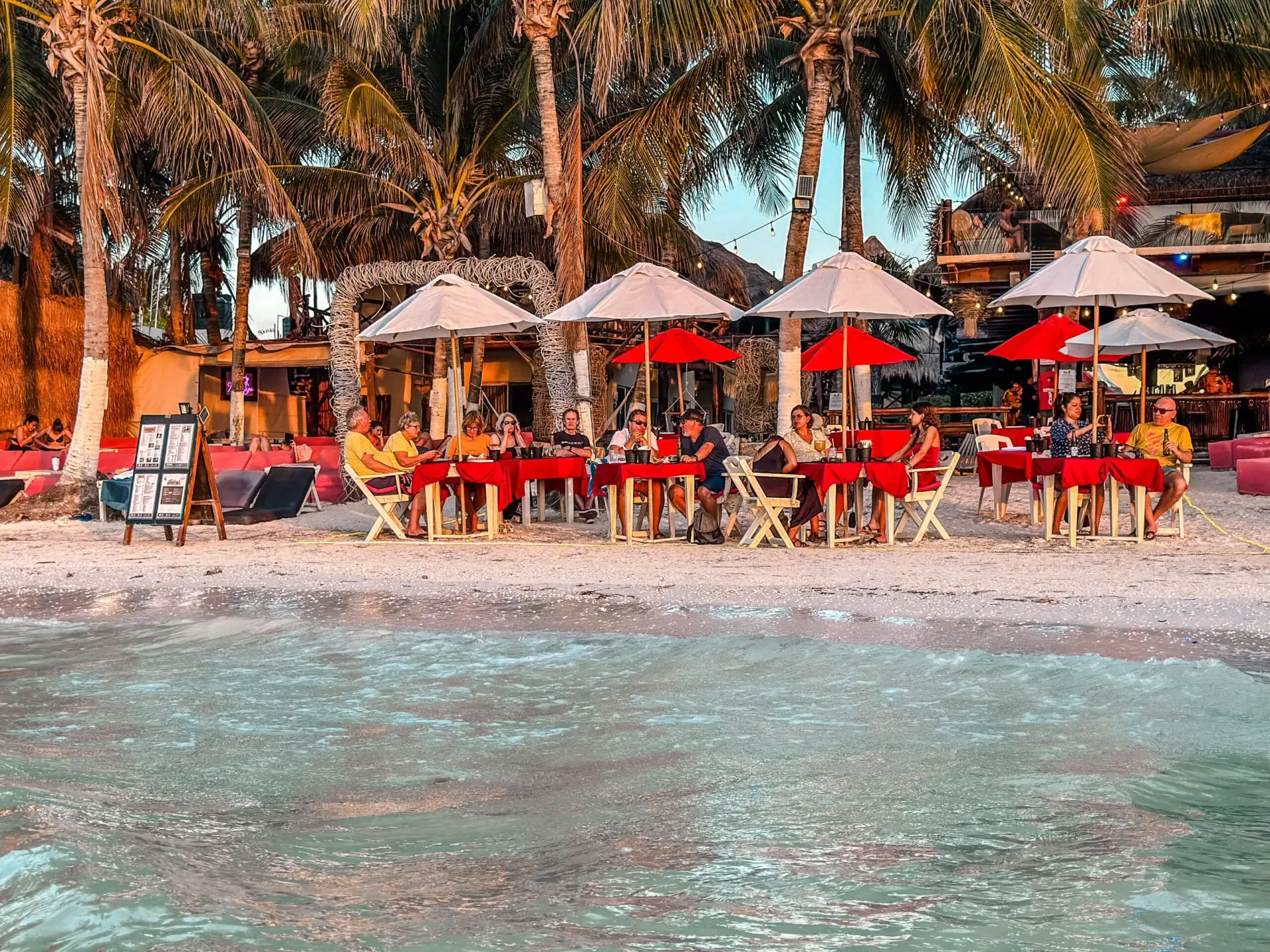 Restaurante junto a la playa con mesas y sombrillas en la arena. Gente cenando, con palmeras al fondo.