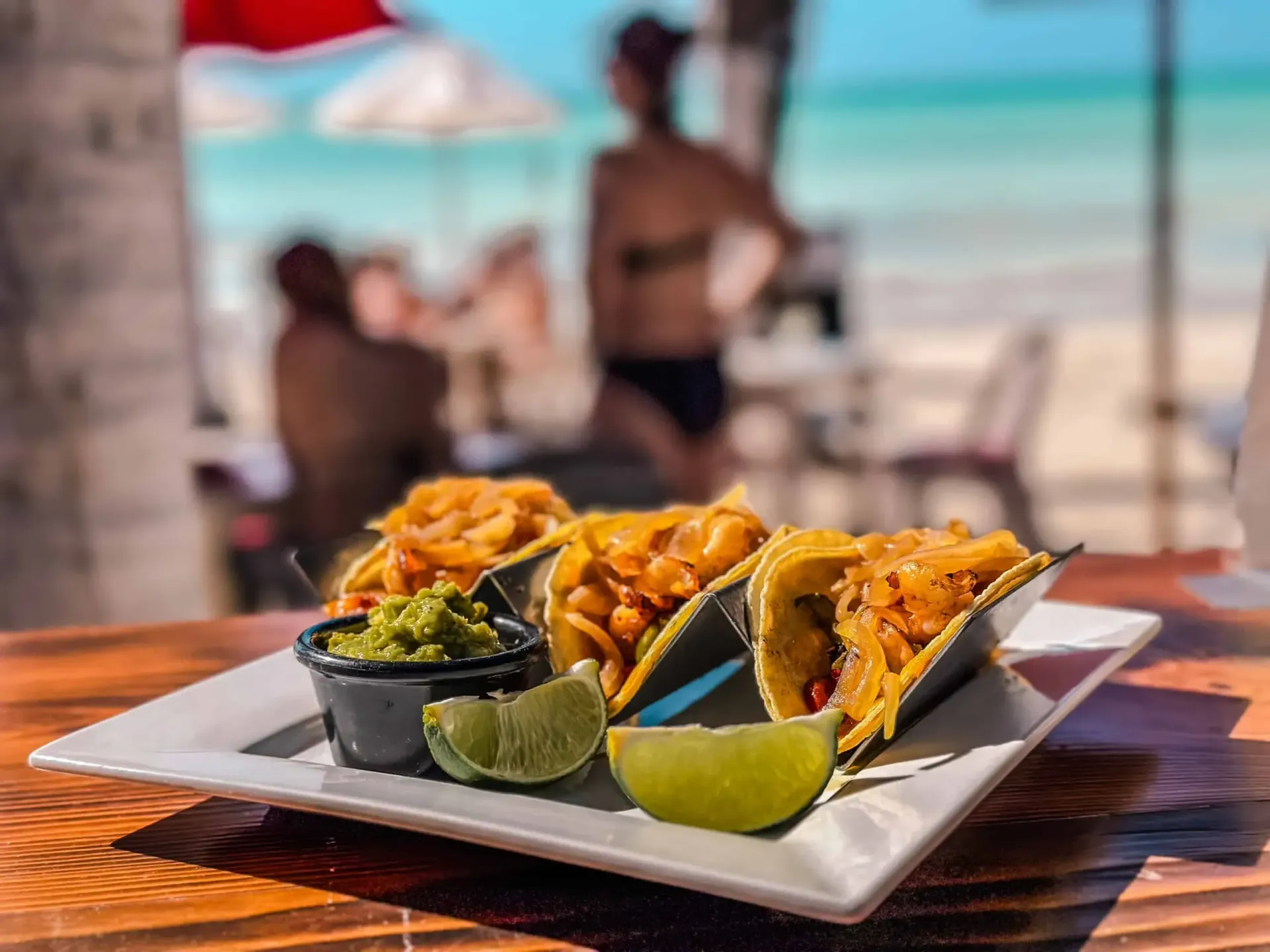 Tacos con guacamole, limón y frijoles en un plato blanco, entorno de playa con gente borrosa.