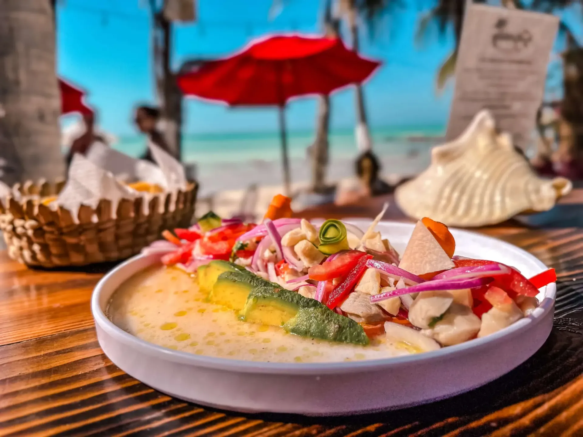 Plato de ceviche en un plato blanco con aguacate, en un restaurante de playa, bajo una sombrilla roja.