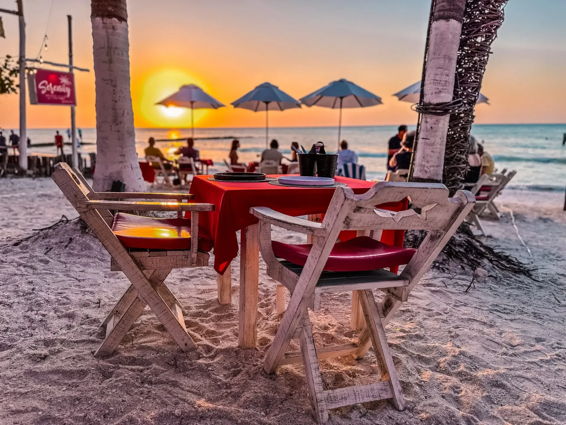 Mesa de playa preparada para cenar con vistas al atardecer. Mantel rojo, sillas blancas y sombrillas.