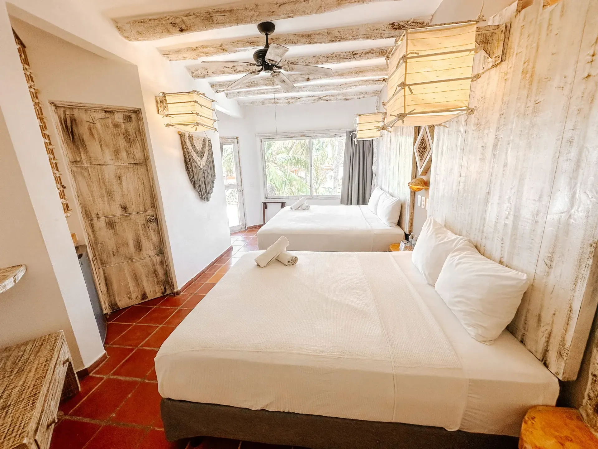 Habitación de hotel luminosa con dos camas, suelo de baldosas rojas, paredes y techo blancos rústicos y ventana grande.