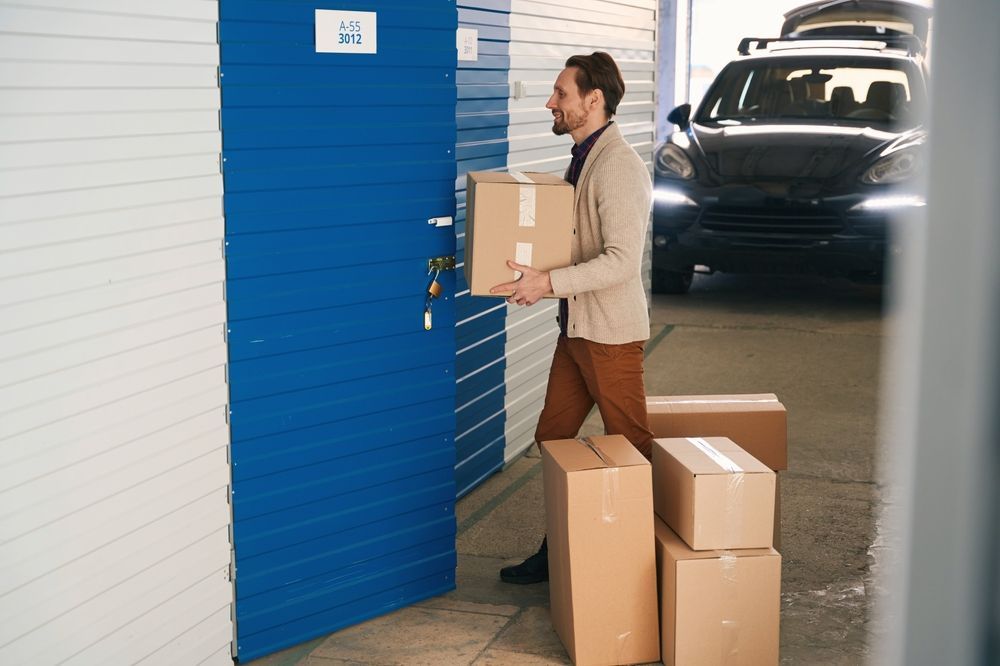 Man carrying a box, entering a blue storage unit; other boxes and a car are present.