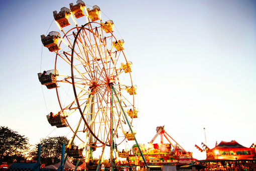 Ferris wheel lit at a fairground at dusk, with colorful amusement rides and tents in the background