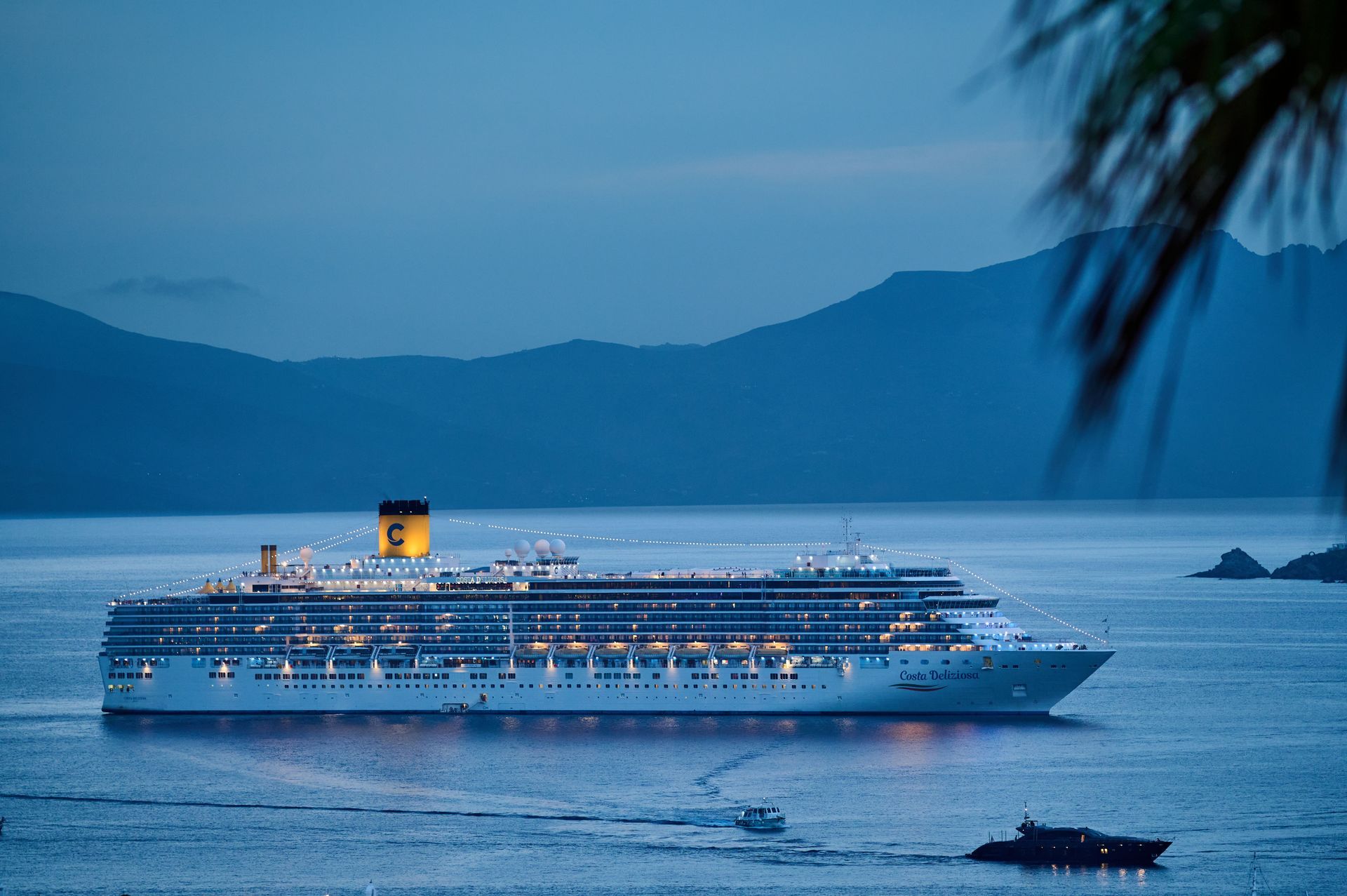 Large cruise ship lit at dusk on calm blue water, with mountains in the background
