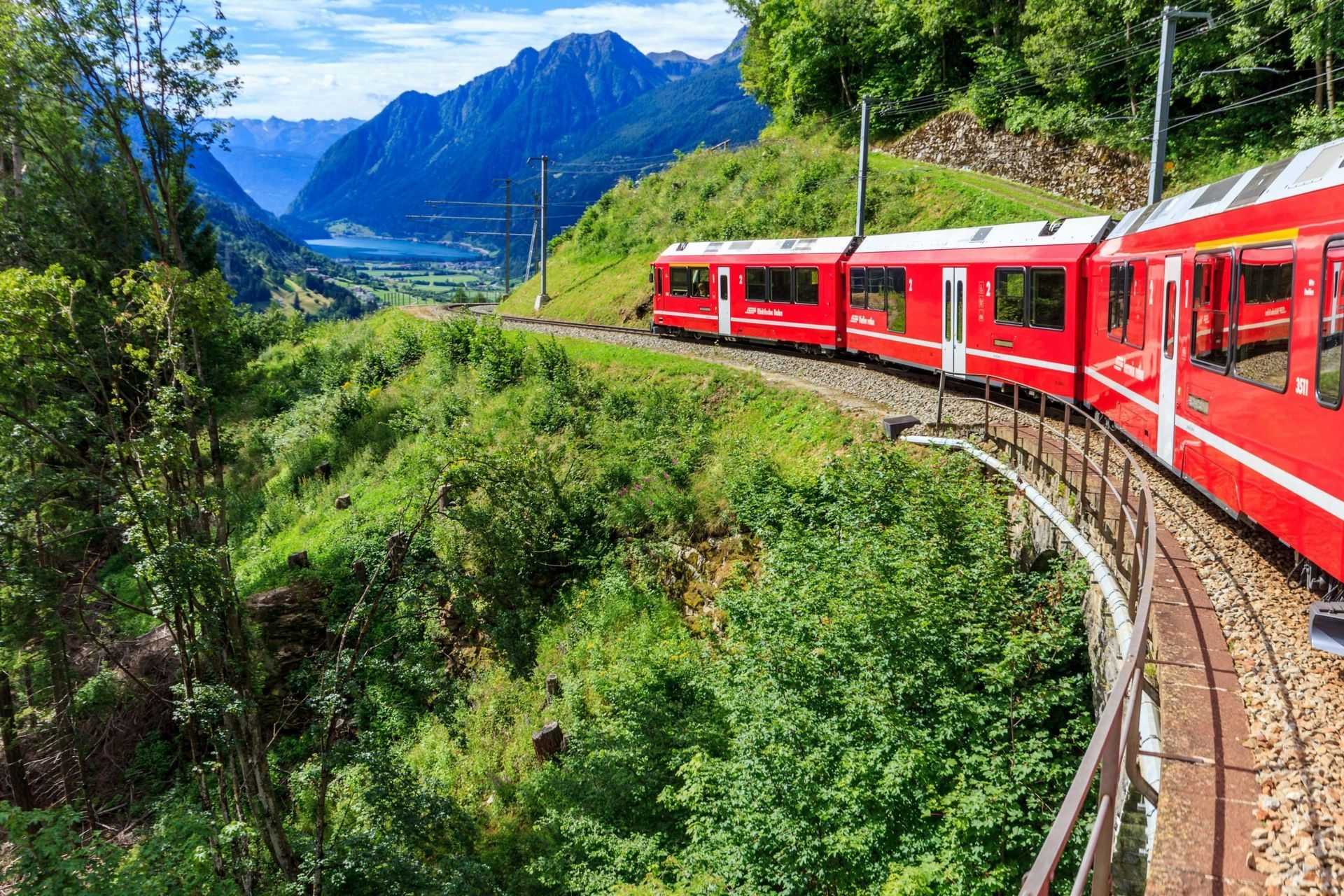 Red train curving through lush green mountains with a blue lake and peaks in the distance