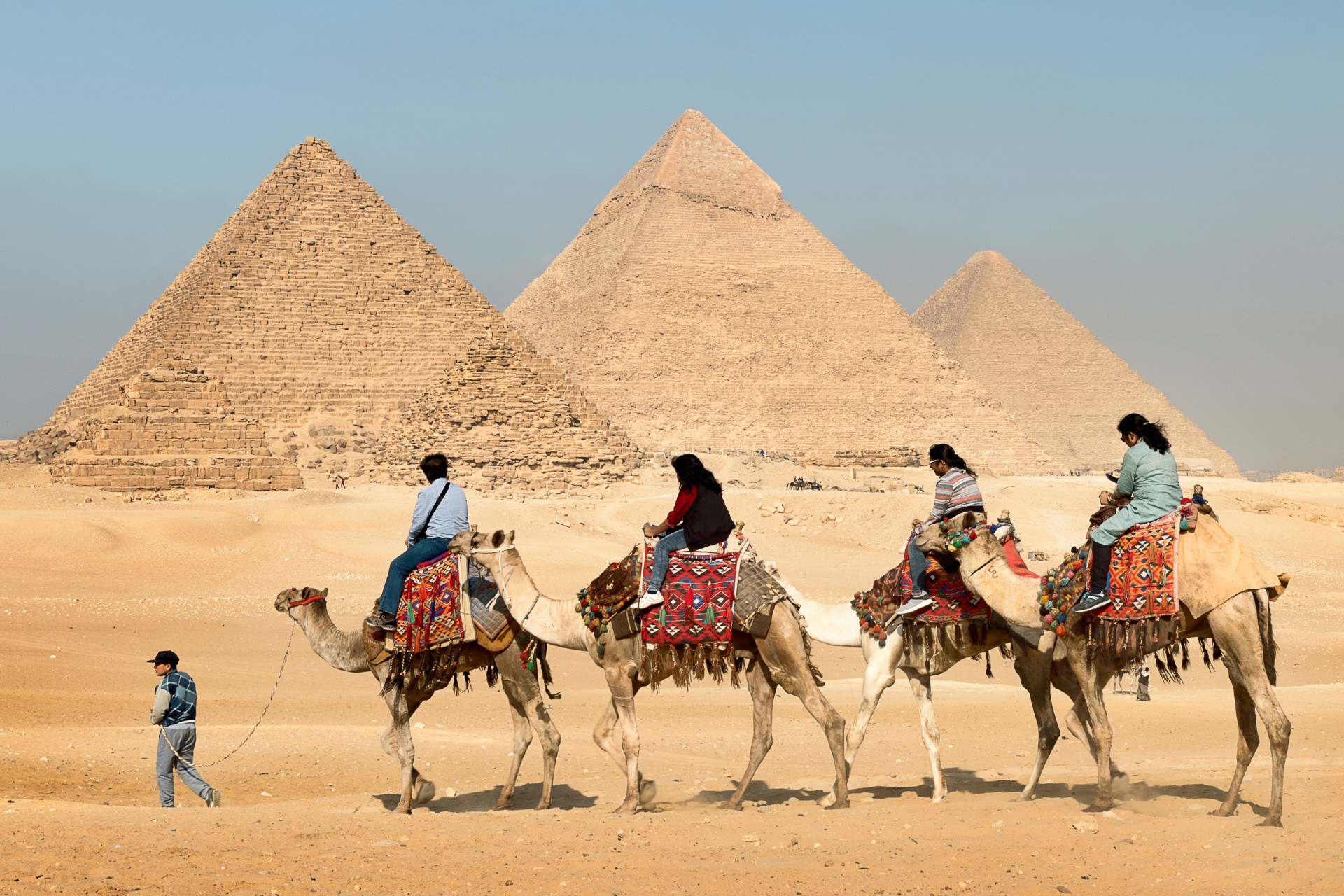 Camels with riders pass before the pyramids in a desert under a clear sky.