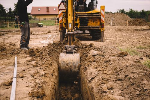 Backhoe digging a trench at a muddy construction site with a worker nearby