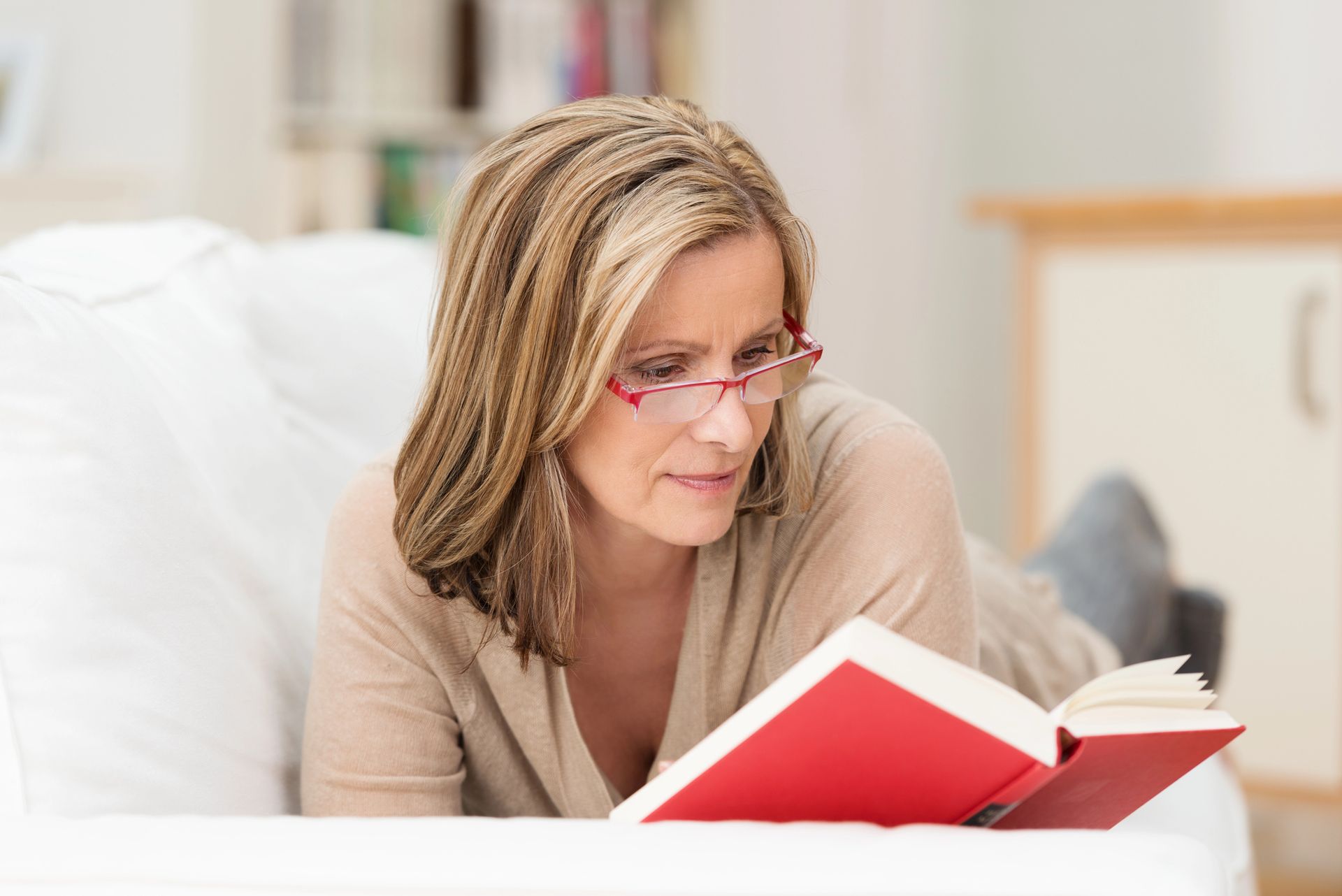 A woman wearing glasses is laying on a couch reading a book.