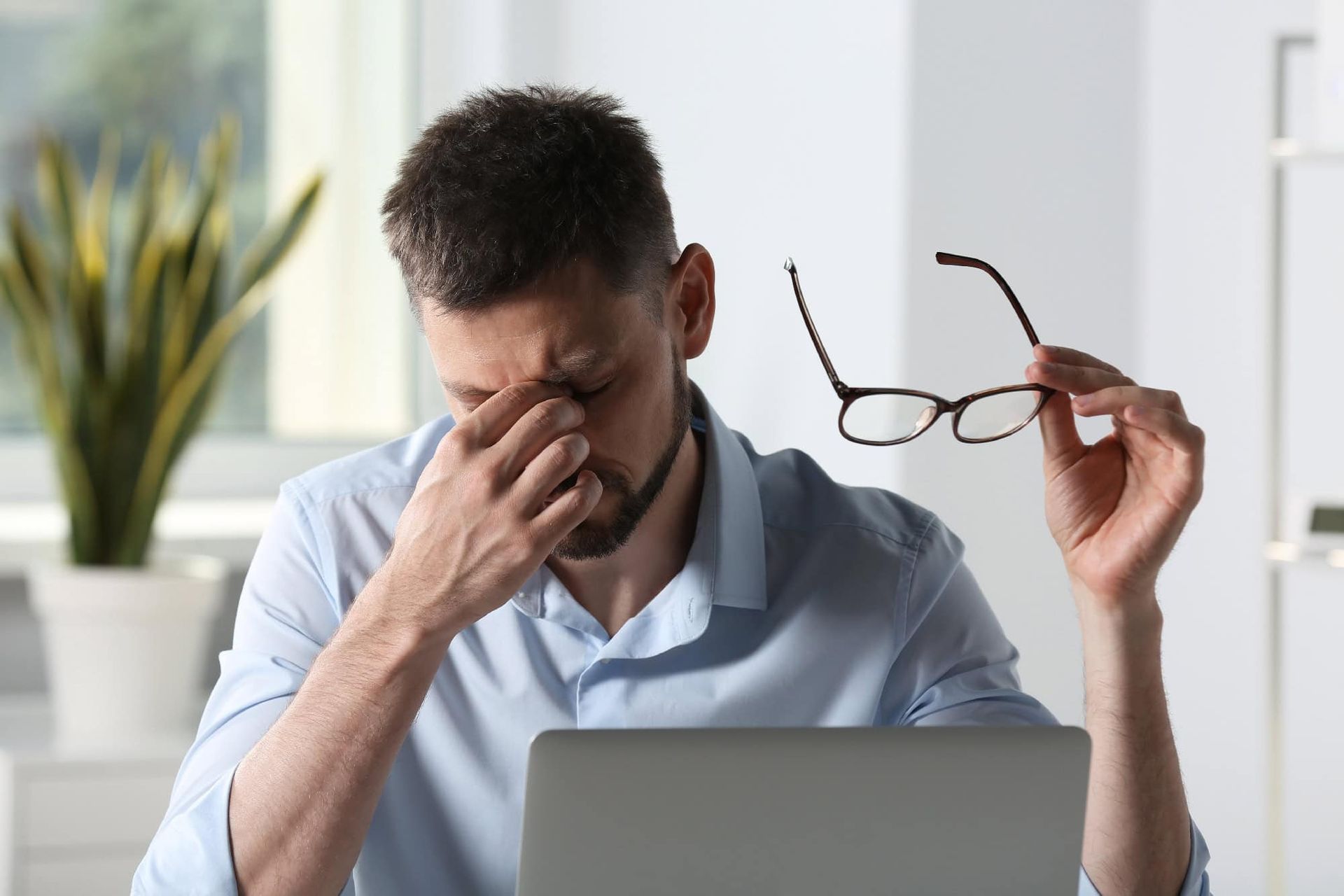 Man rubbing eyes beside a laptop while holding eyeglasses in a bright office