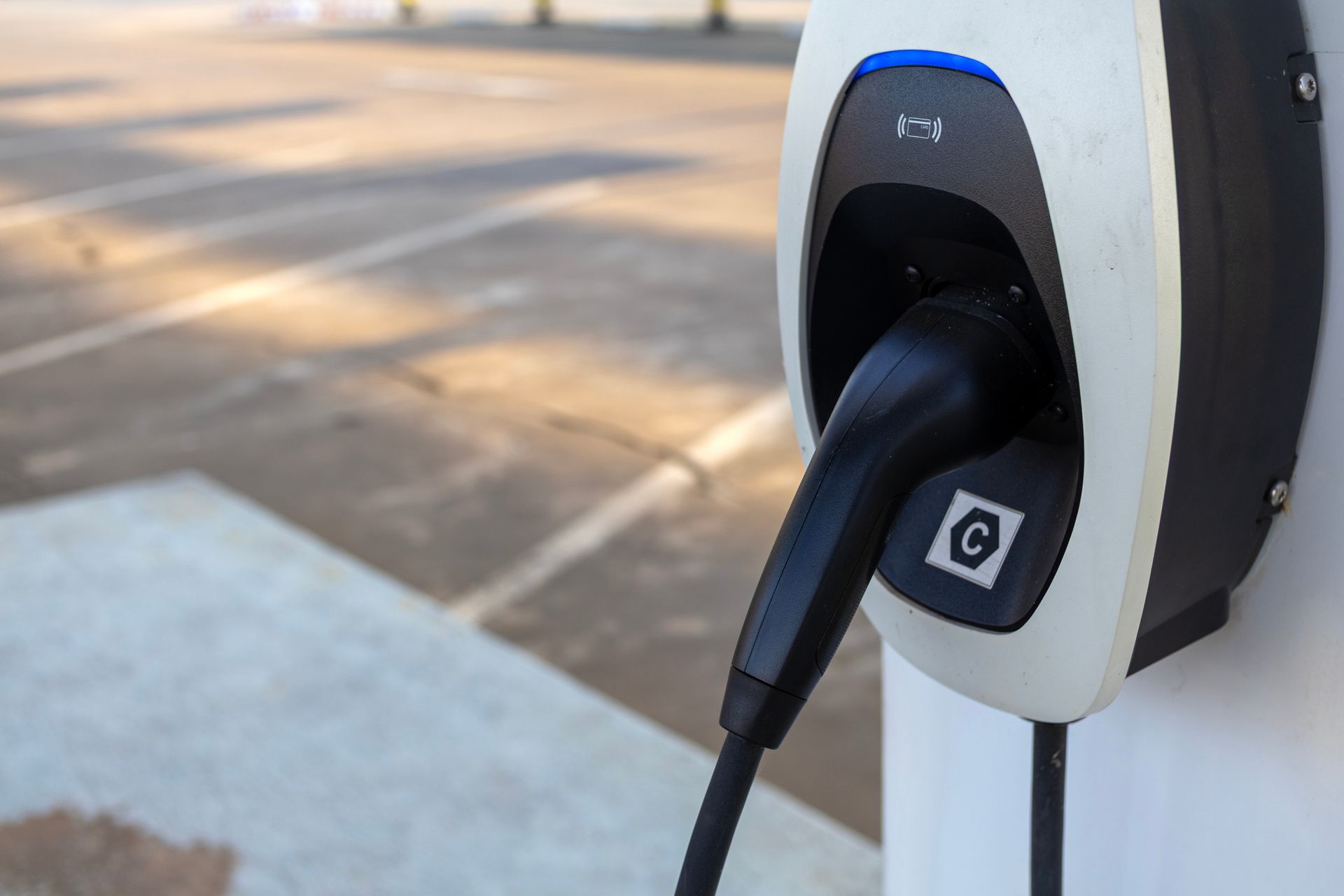Electric vehicle charger with a black charging cable plugged in; mounted on a white and black station in a parking area.