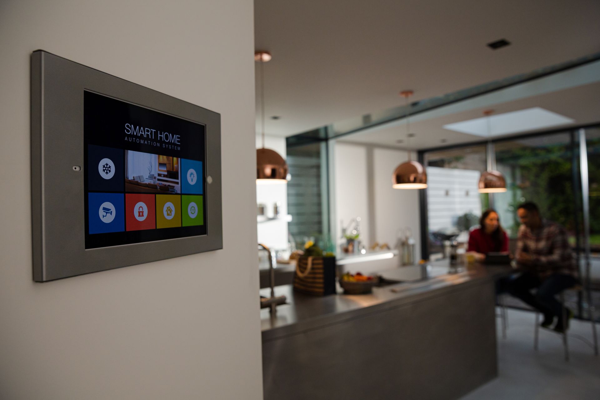 Wall-mounted smart home control panel with a touchscreen displaying a colorful interface. Two people are blurred in the background.