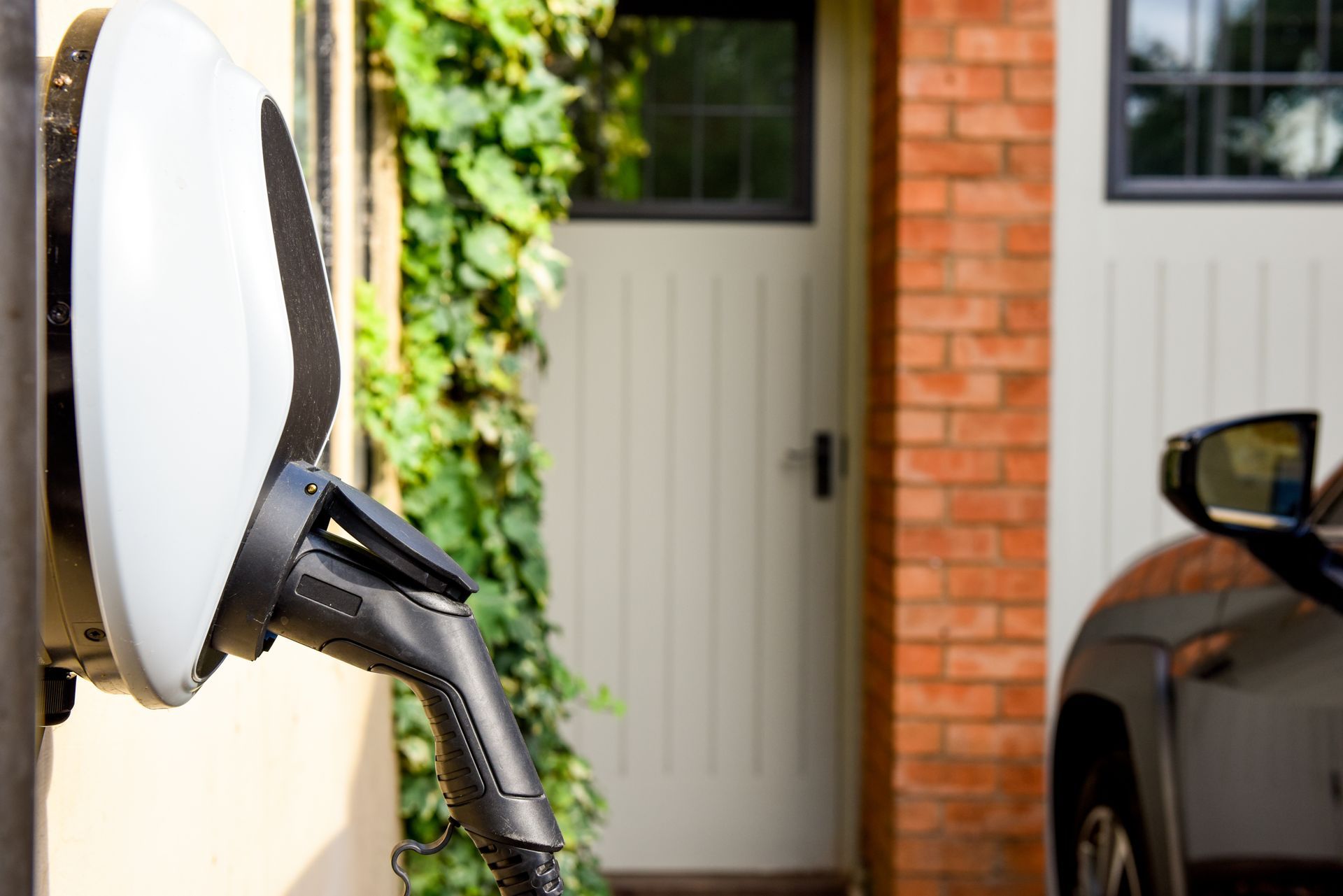 Electric vehicle charging station on a beige wall, with a charging cable connected to a car parked beside a brick house.