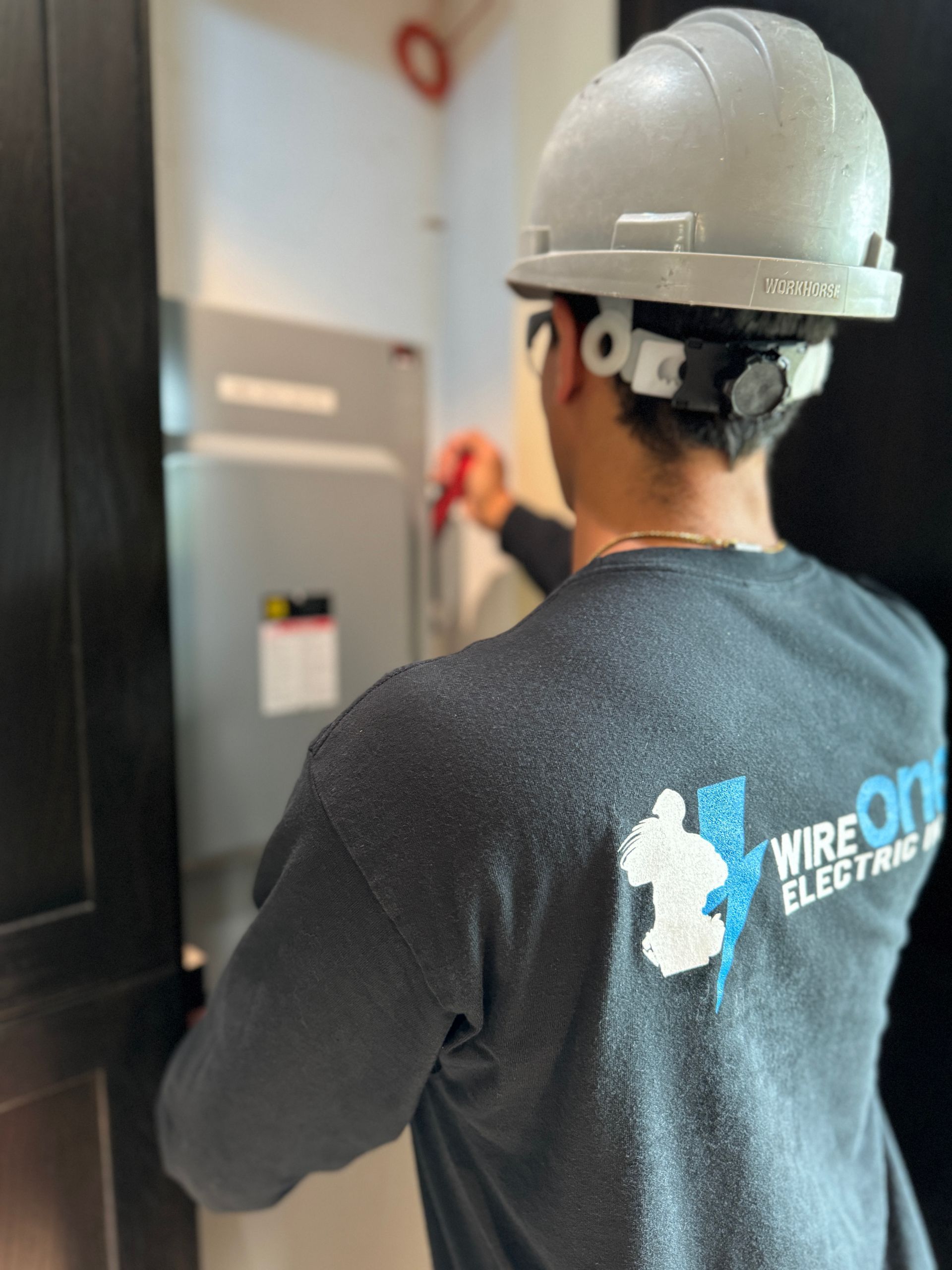 Electrician in hard hat working on an electrical panel. He's wearing a gray shirt with the WireOne Electric logo.