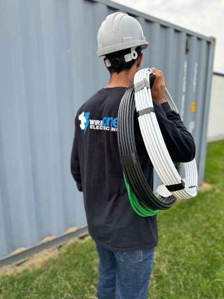 A construction worker in a hard hat carries bundled electrical wires outside near a storage container. The wires are white, black, and green.