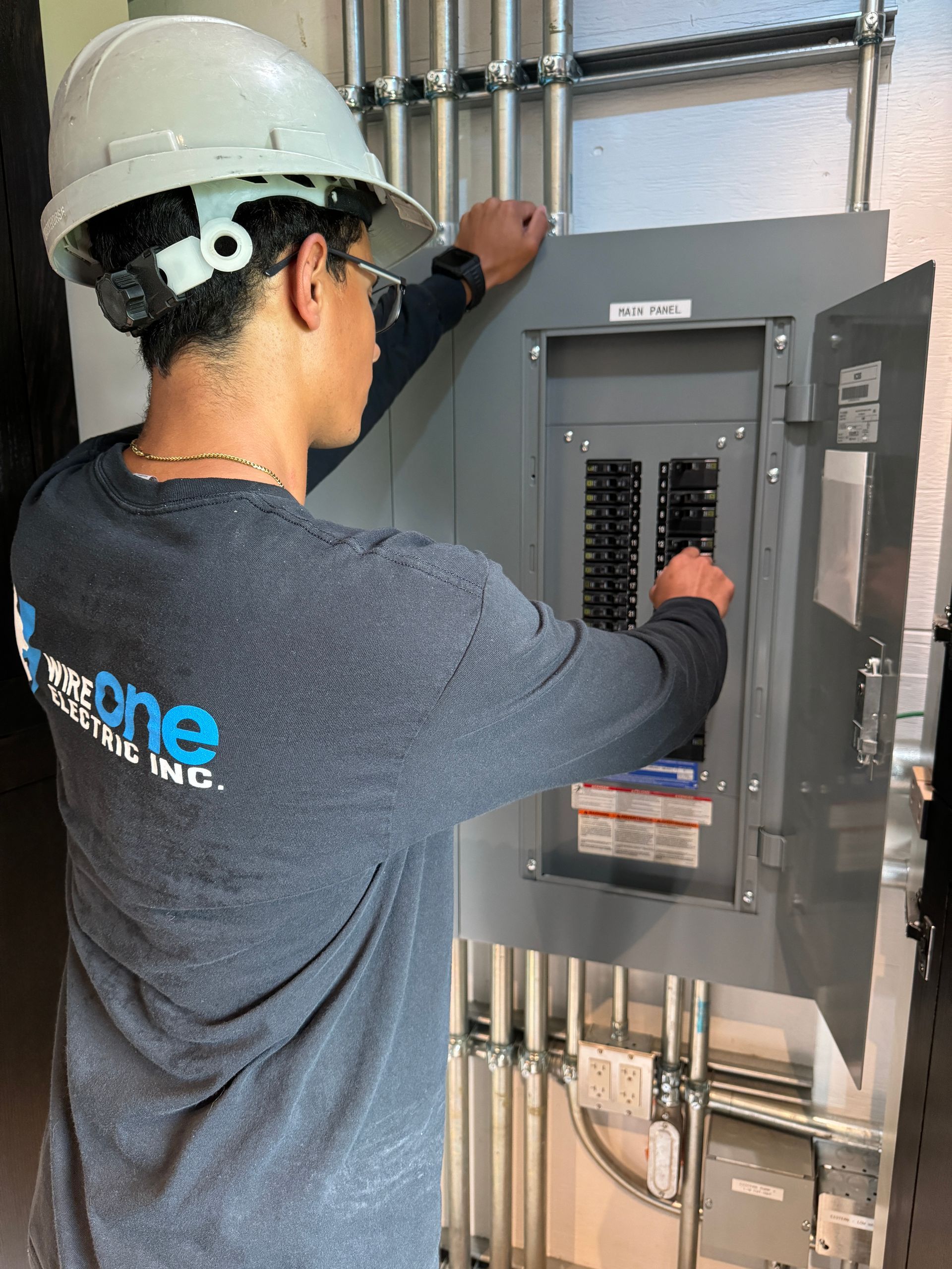 A person in a hard hat and long-sleeve shirt working on an electrical panel. They are turning a circuit breaker.