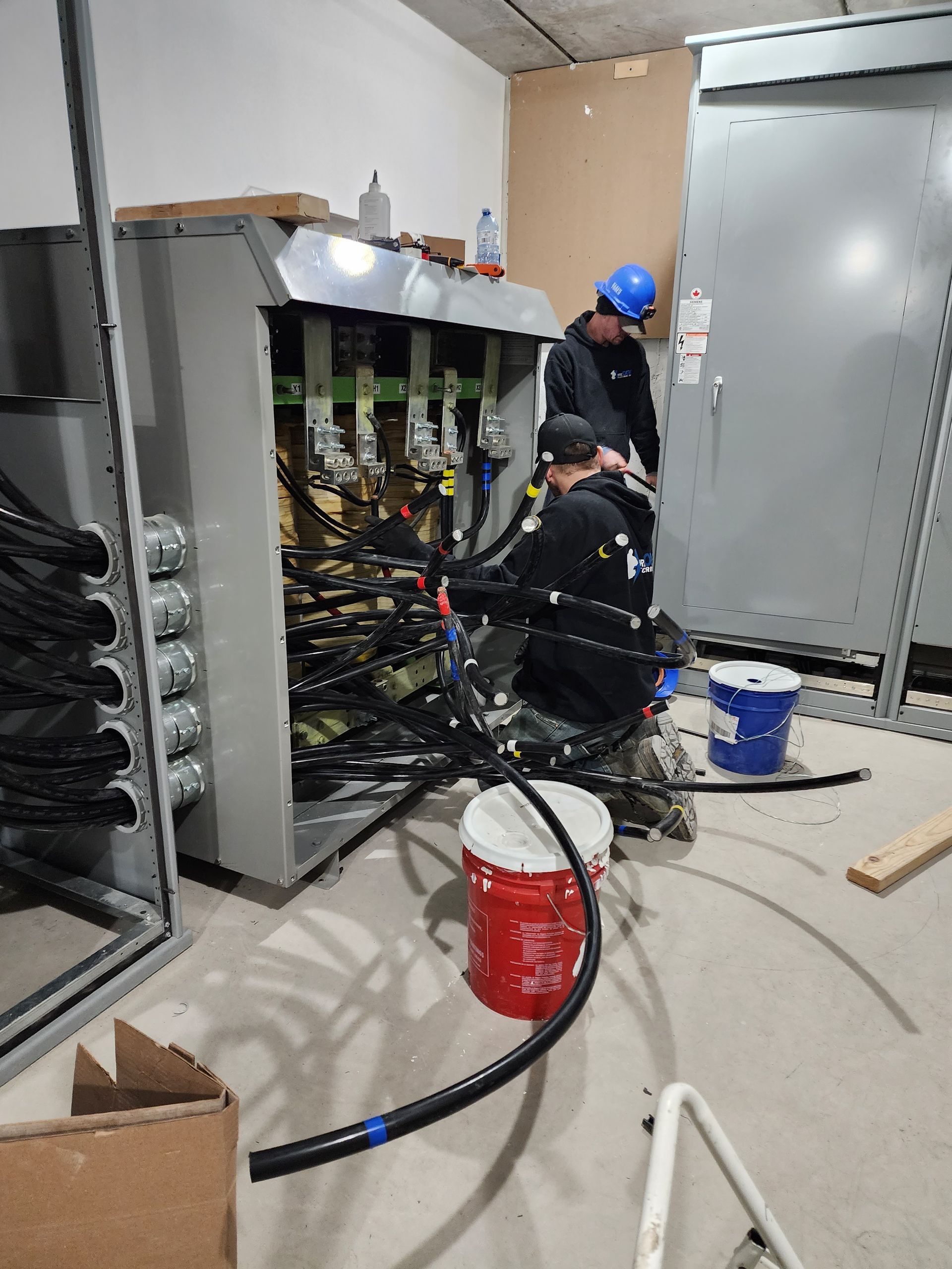 Two electricians working inside an electrical panel in a construction setting. Wires and connections are visible, along with tools and a paint bucket.