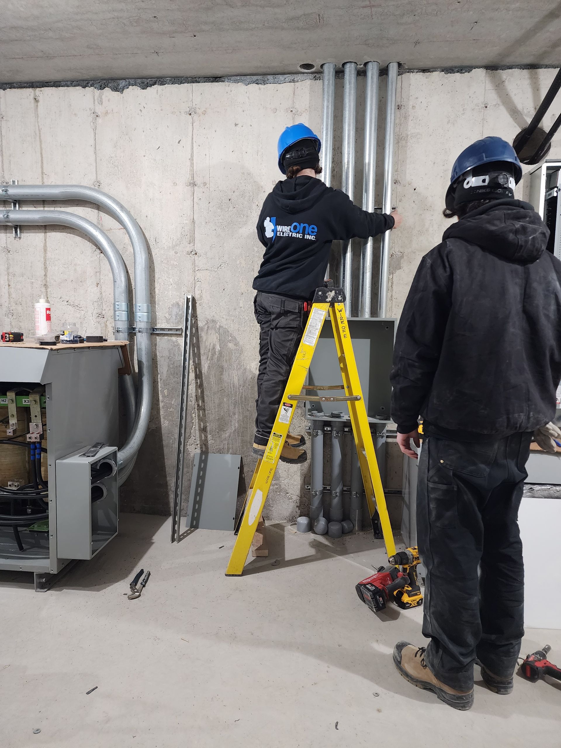 Two electricians in a construction setting. One on a yellow ladder works on conduit, the other stands nearby. Concrete walls and equipment are in the background.