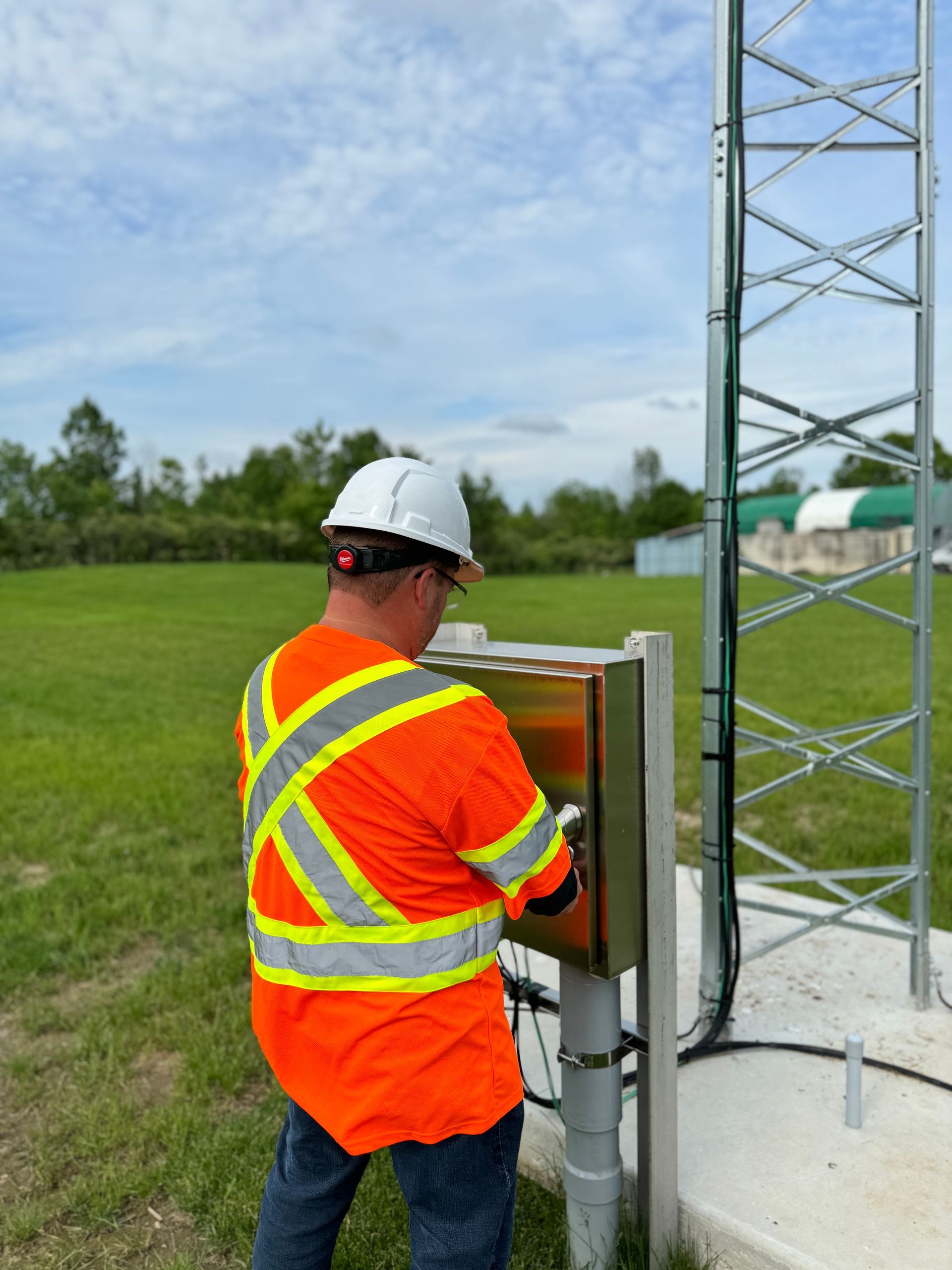 A worker in an orange safety vest and hard hat inspects a metal box at the base of a communication tower on a grassy field.
