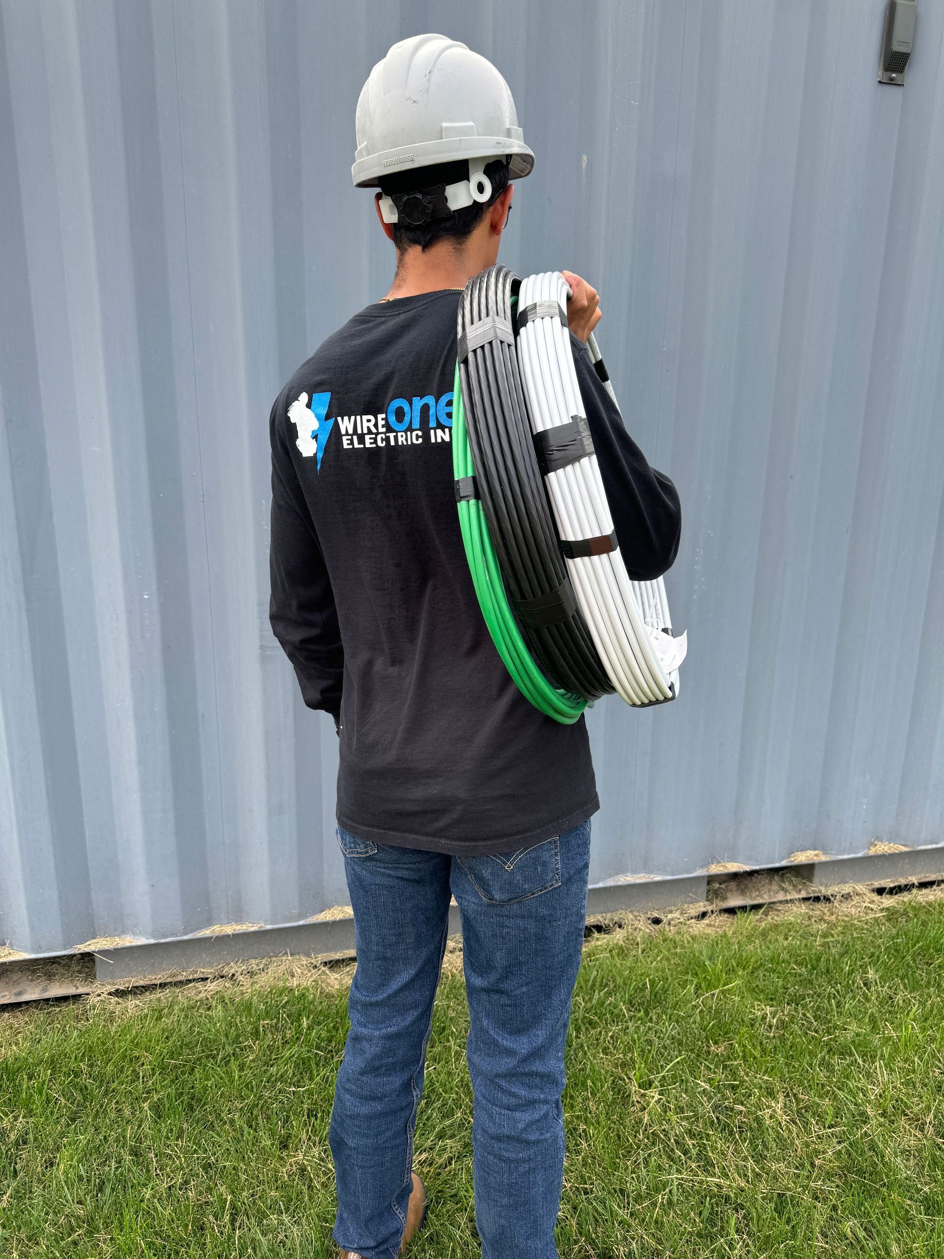 Person in a hard hat carrying a large coiled cable, standing by a corrugated metal wall.