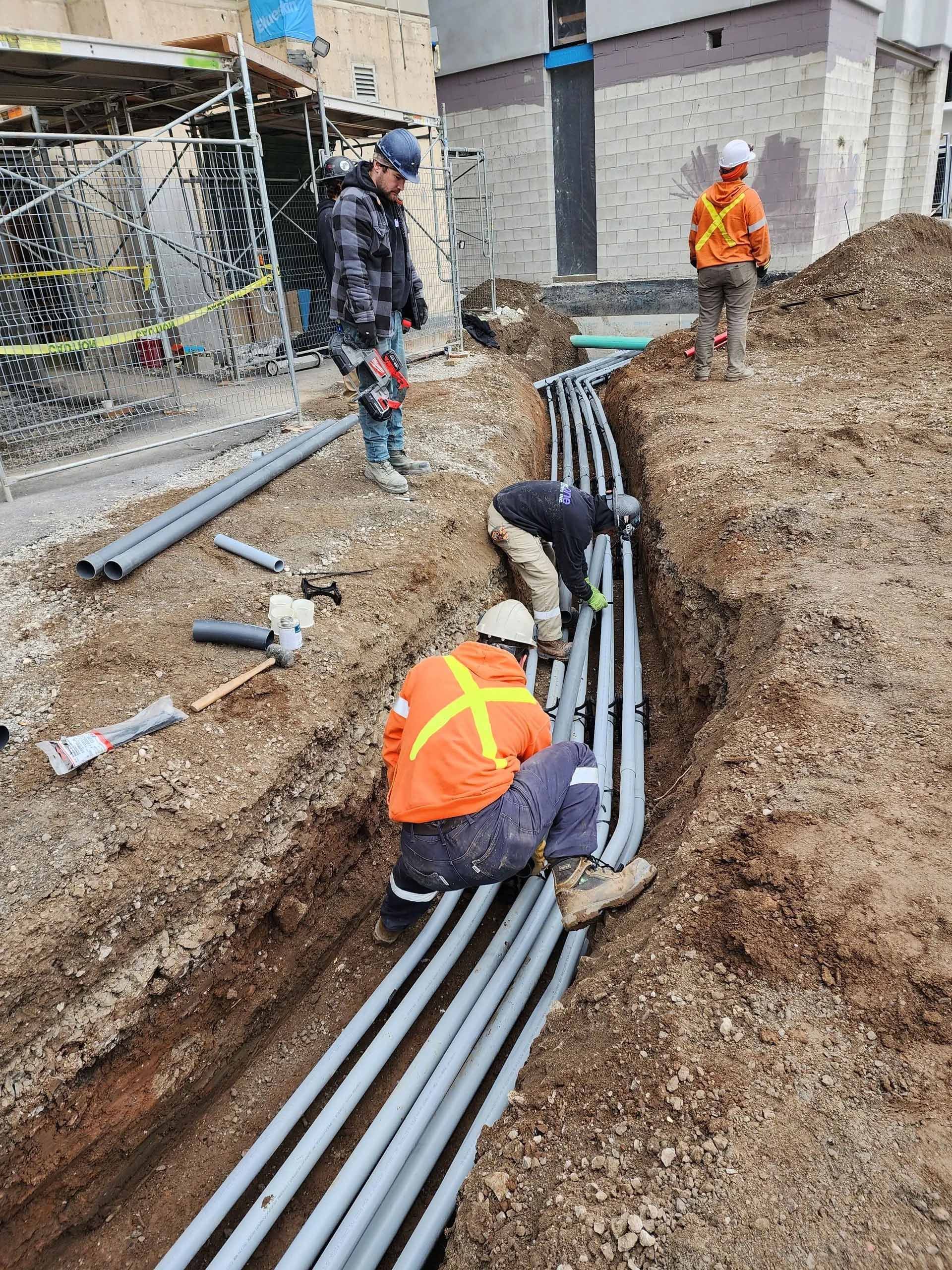 Construction workers laying gray pipes in a trench beside buildings at an excavation site