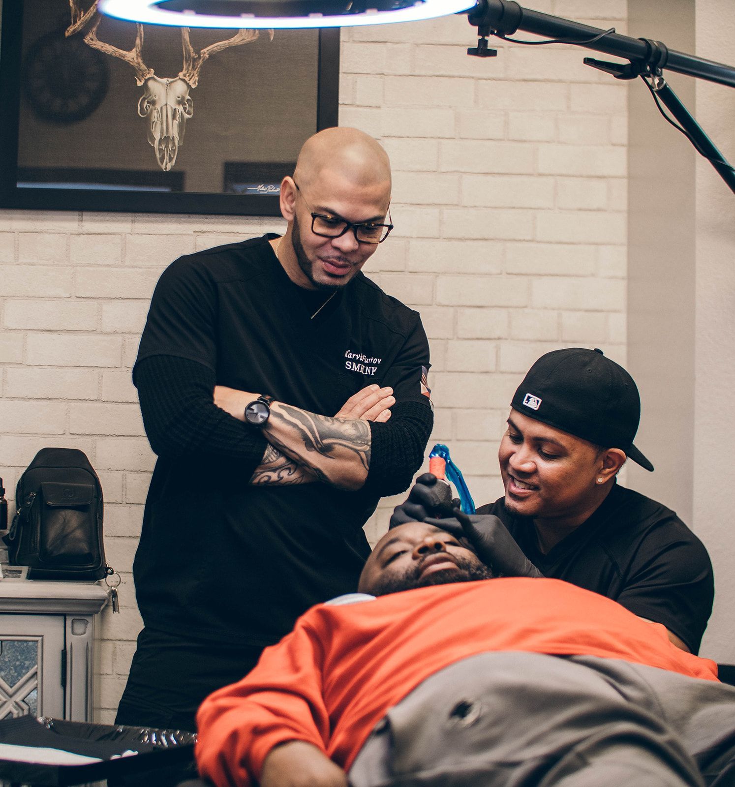 Two people in a tattoo studio work on a client lying on a chair under a hanging lamp.