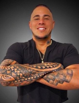 Smiling person in a black shirt with crossed tattooed arms against a gray background