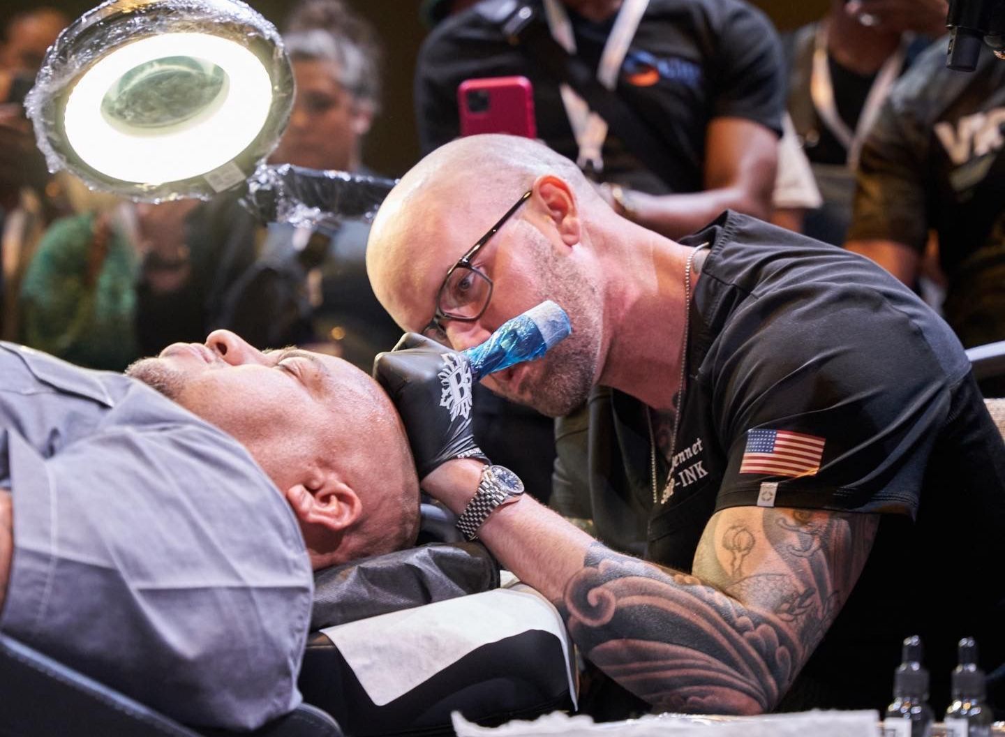 Tattooed man tending to a reclining person in a crowded event setting, focused and close up