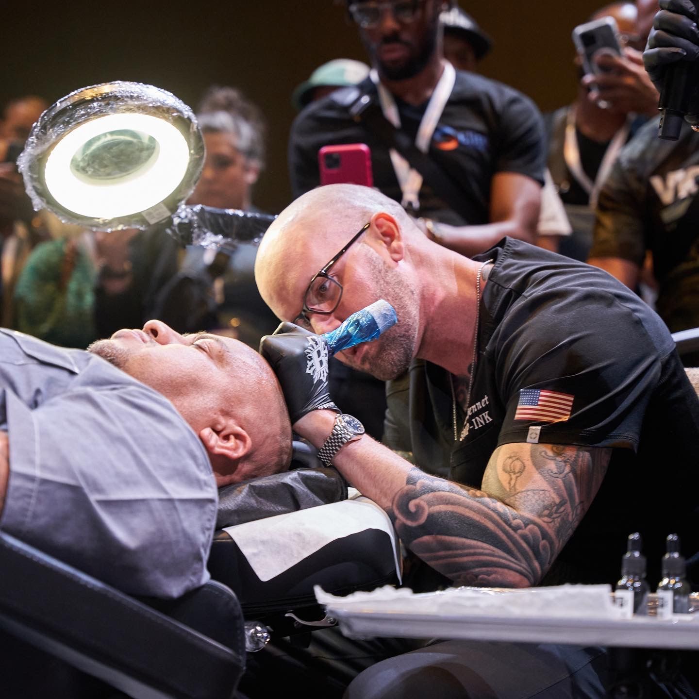Tattoo artist working on a client's arm at a live event, with a bright ring light and onlookers nearby
