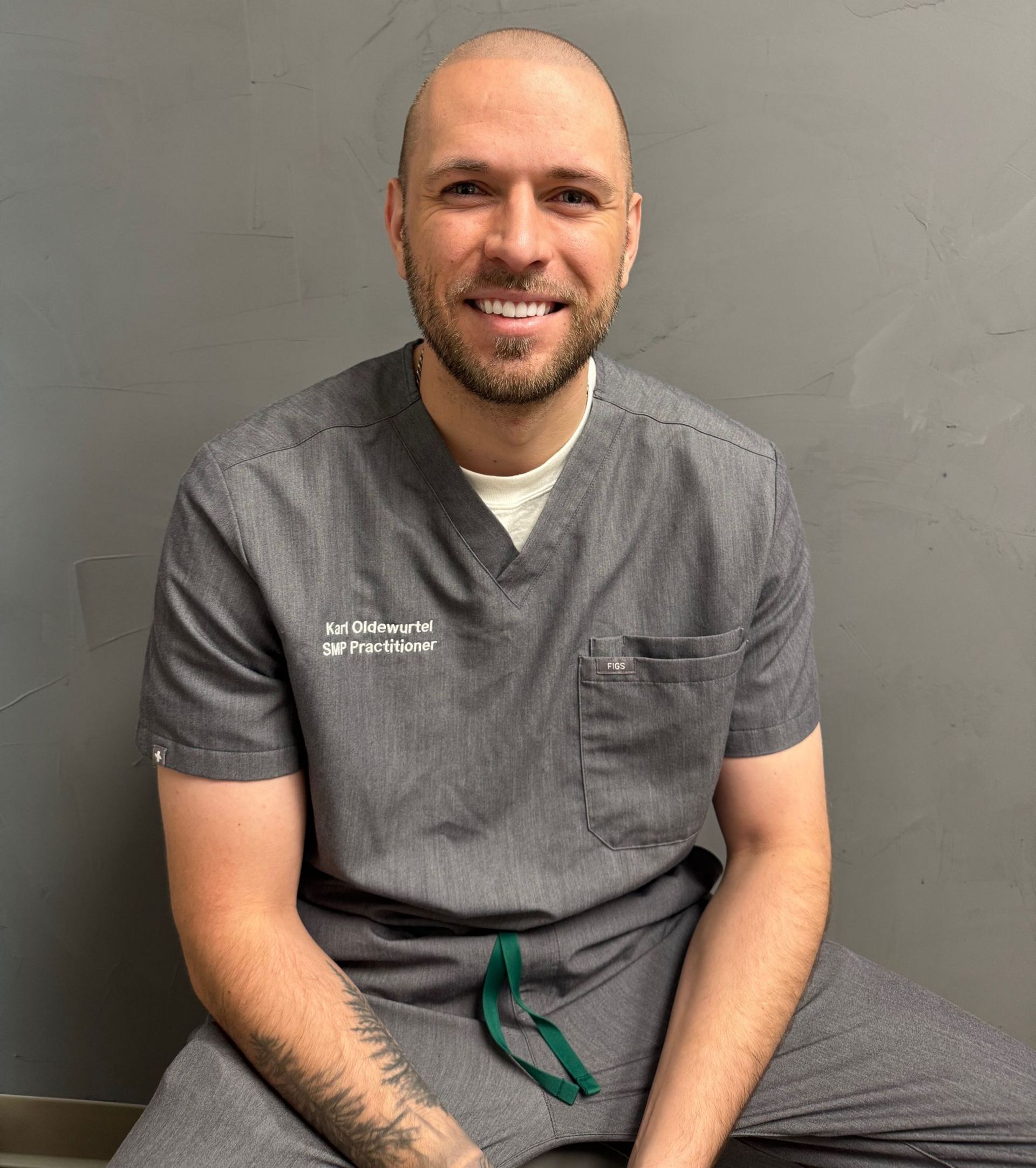 Smiling bald person in gray scrubs seated against a gray wall, with tattoos on one forearm