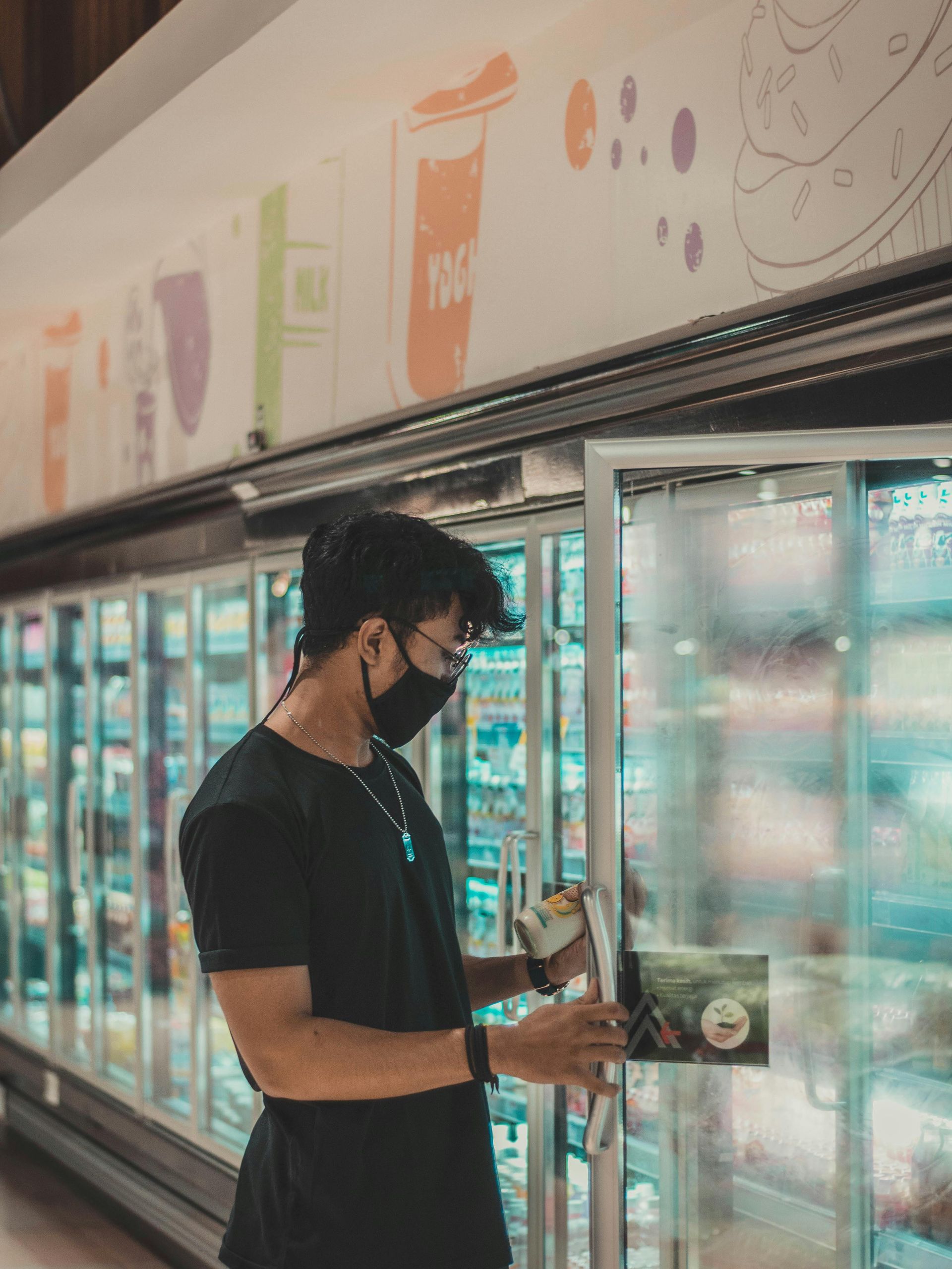 A man wearing a mask is standing in front of a refrigerator in a grocery store.