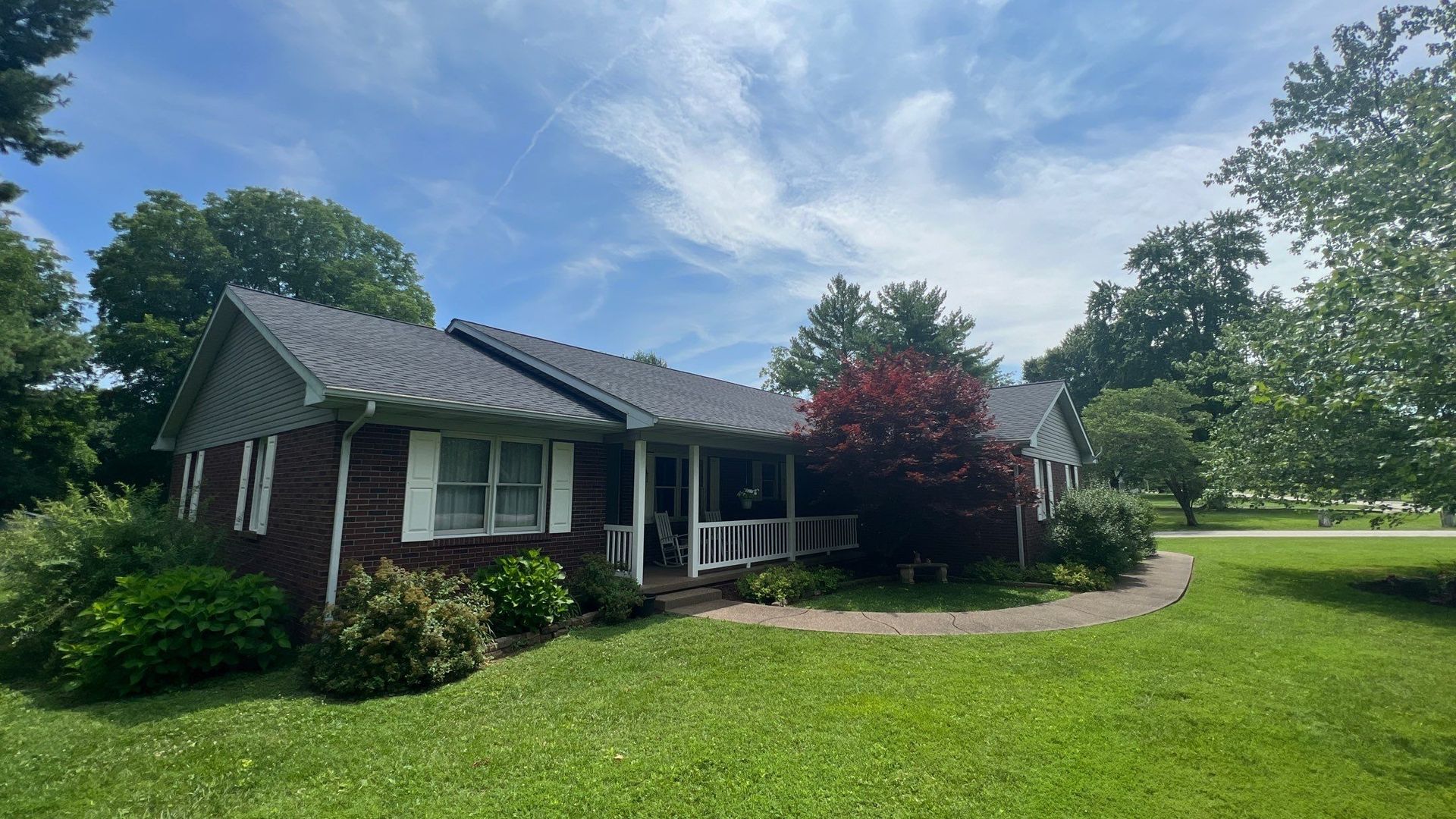 A brick house with a porch and a gray roof is sitting on top of a lush green field.