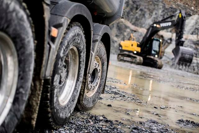Pneumatici di camion su terreno fangoso con un escavatore giallo sullo sfondo, probabilmente un cantiere edile.