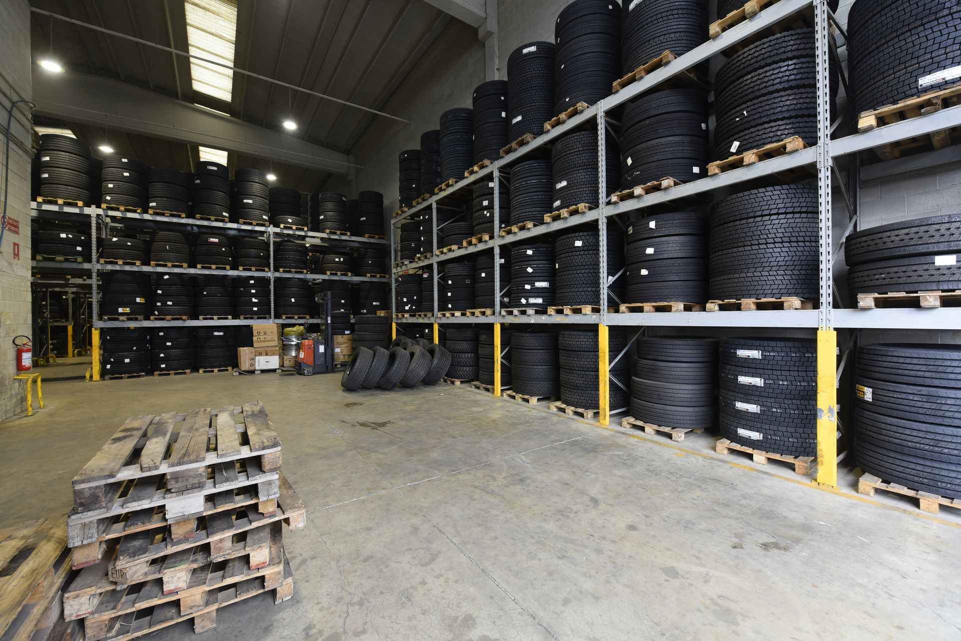 Interno di un magazzino con scaffali pieni di pneumatici accatastati. Pallet in primo piano e un estintore sulla parete.