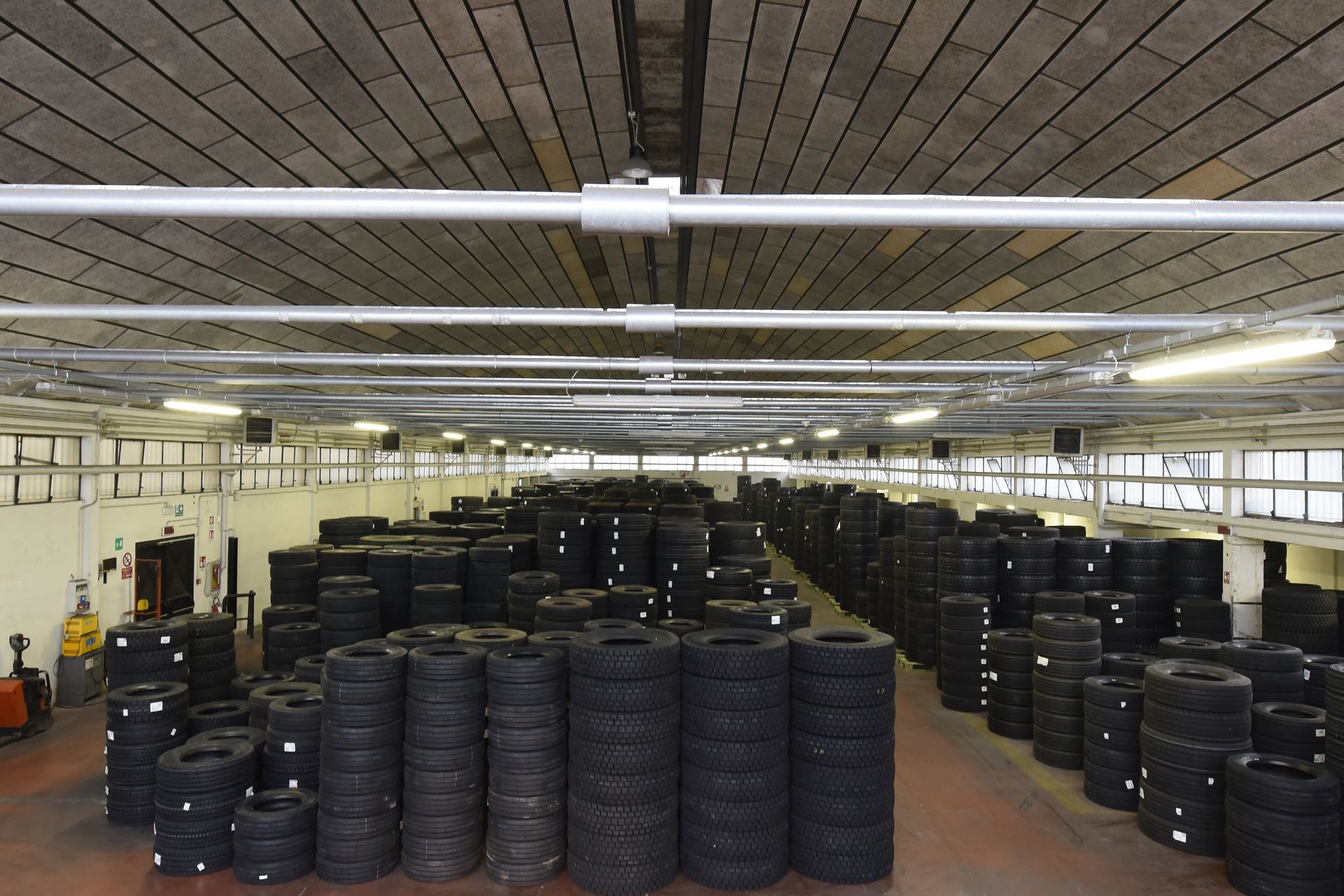 Magazzino pieno di pneumatici accatastati, sotto un soffitto industriale con illuminazione dall'alto.