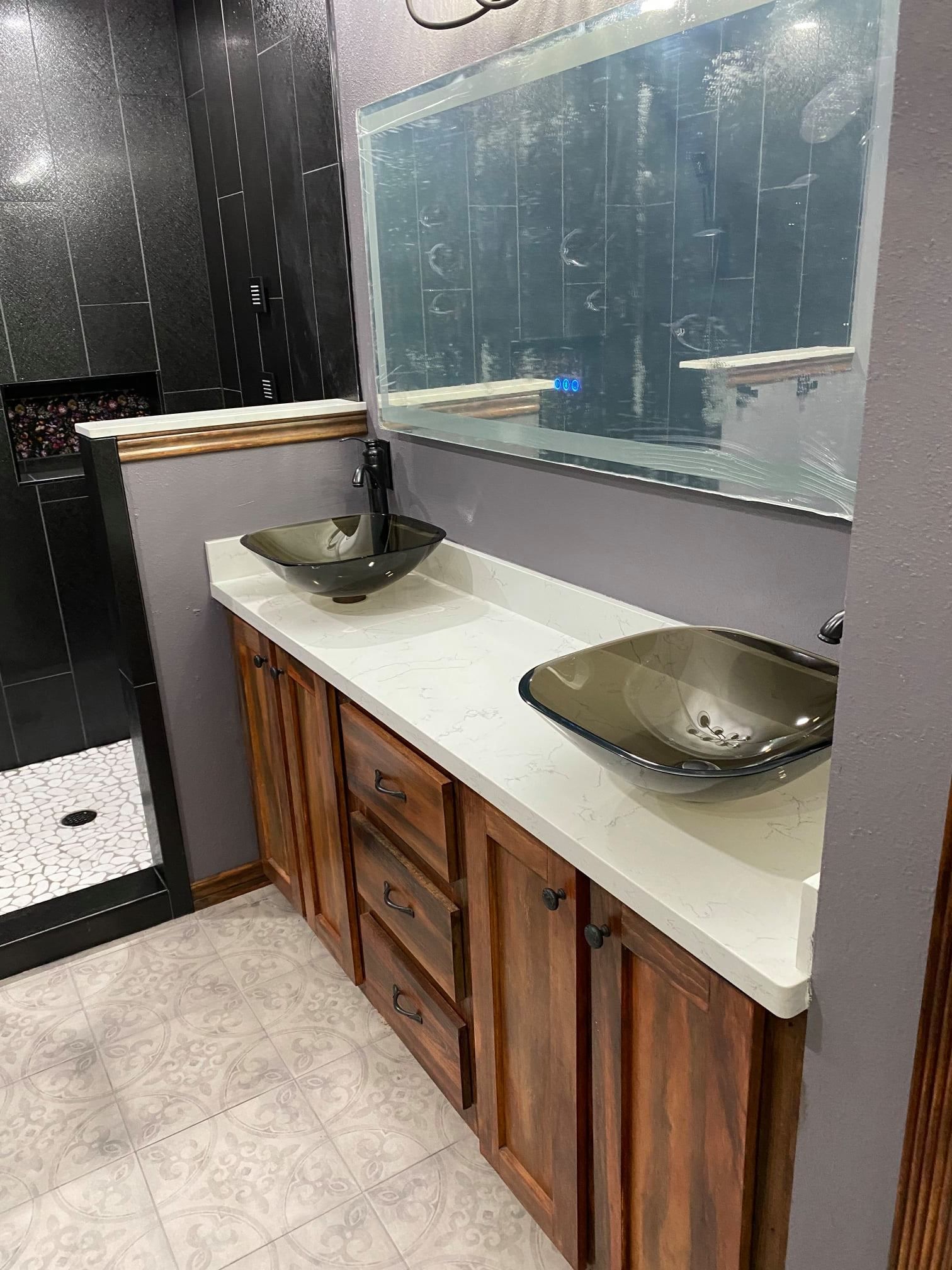 Bathroom vanity with two black vessel sinks, white countertop, and large mirror on a dark-tiled wall.
