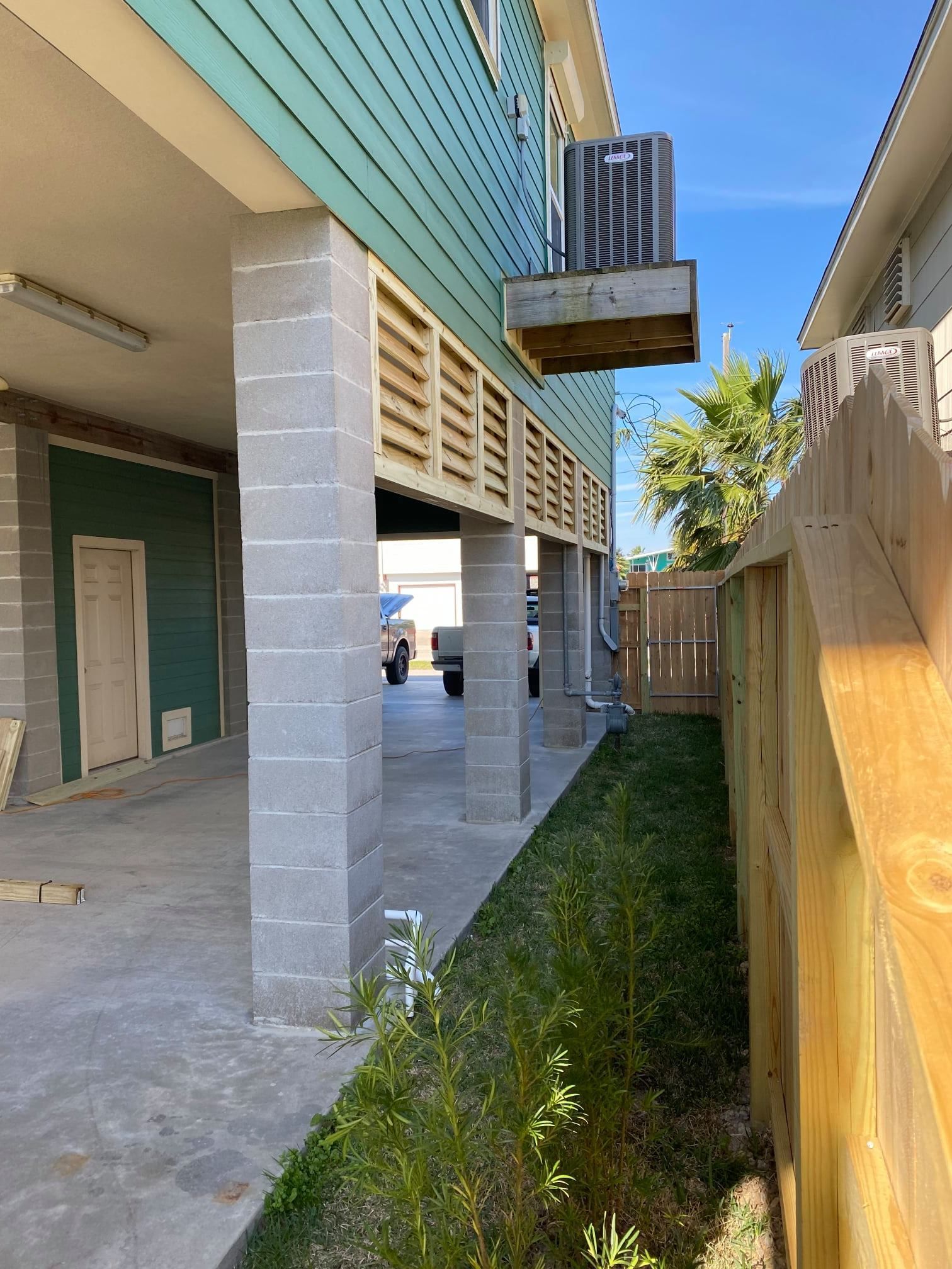 Outdoor walkway beside a building, with concrete columns, grass strip, wooden fence, and clear blue sky.