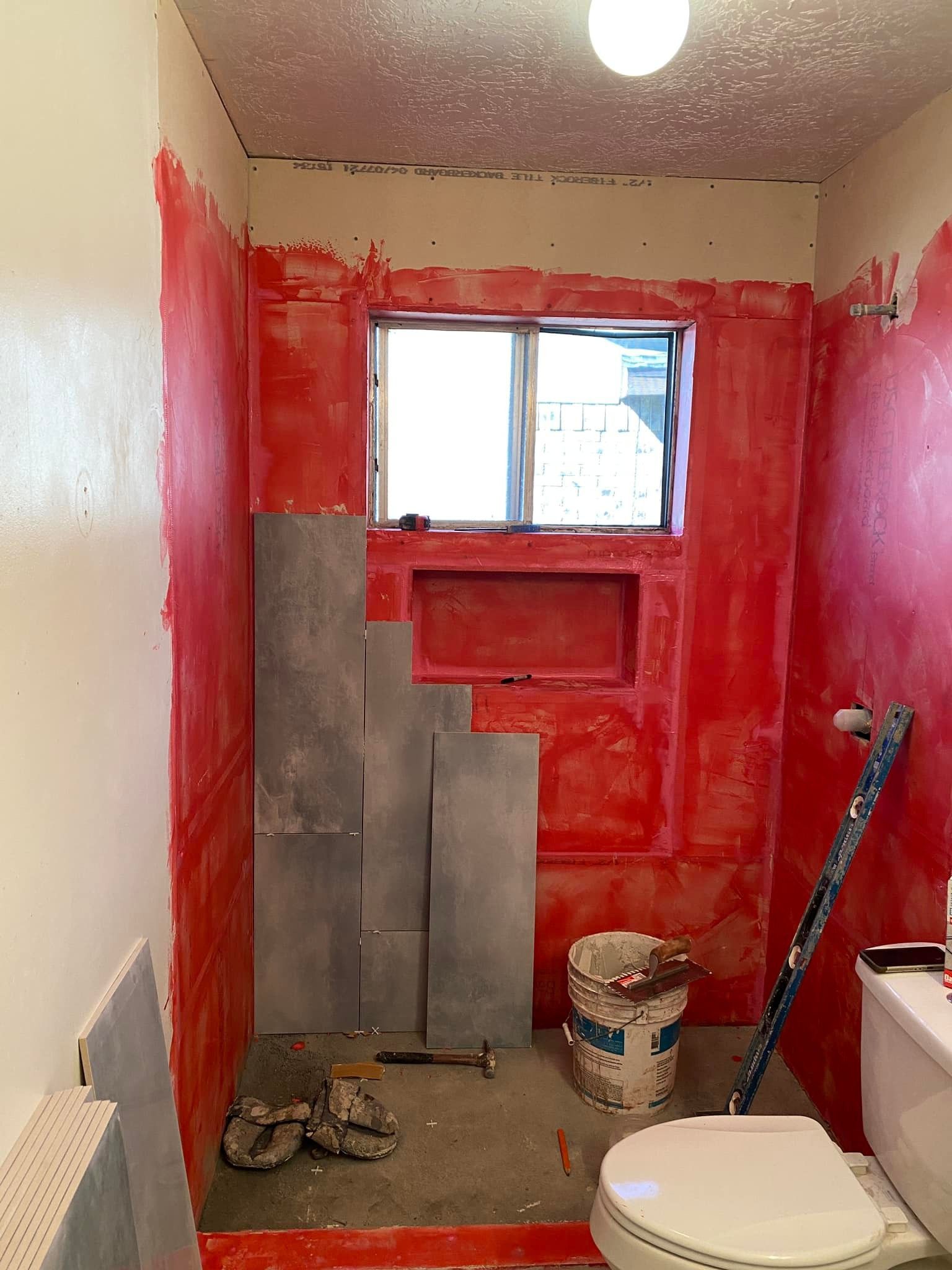 Partially renovated bathroom with red painted walls, gray tile, window, toilet, and cleaning supplies
