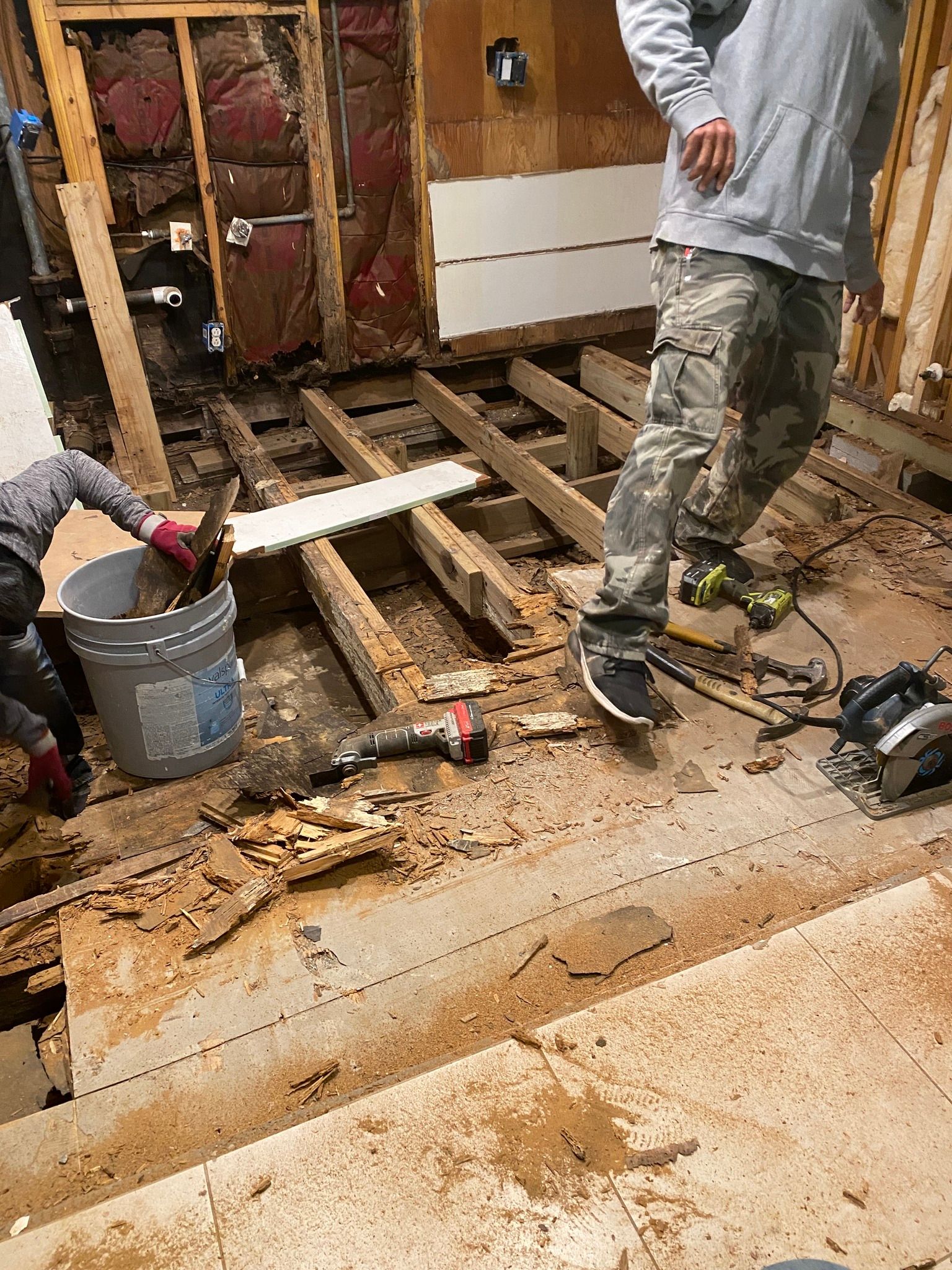 Two workers amid a floor demolition, with exposed joists, tools, debris, and a gray bucket.