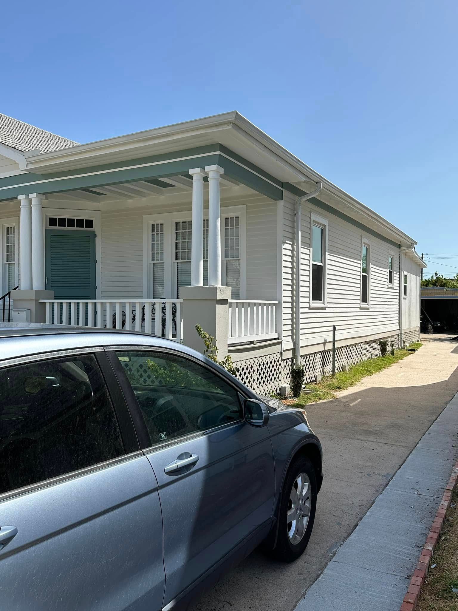 White house porch beside a parked dark sedan on a sunny street, with a sidewalk and blue sky.