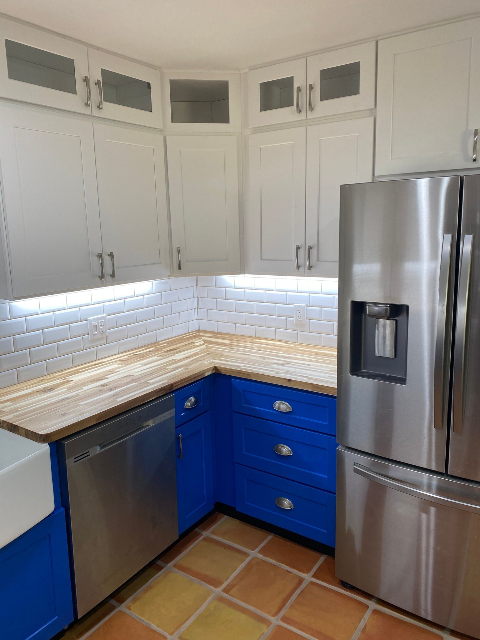 Modern kitchen corner with white cabinets, blue lower drawers, stainless dishwasher and refrigerator, tiled floor.