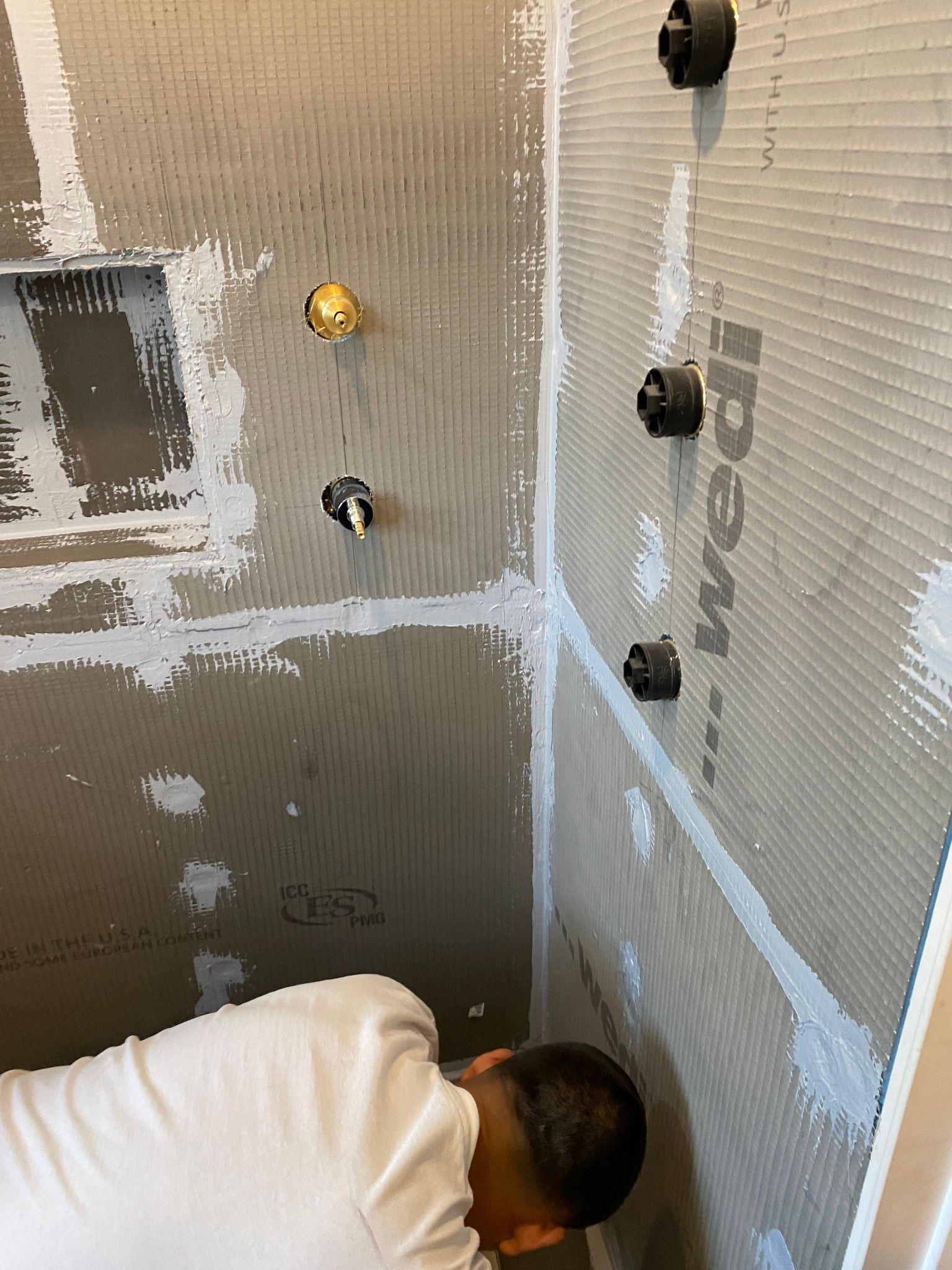 Person installing shower wall tile with a notched trowel in a bathroom corner