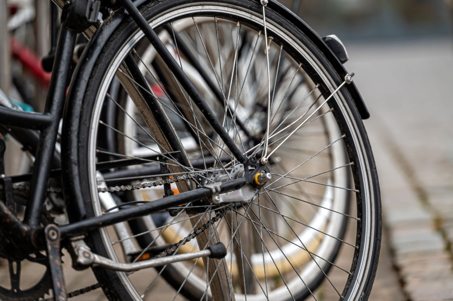 A close-up, side view of a black bicycle rear wheel with thin silver spokes parked on a blurred city sidewalk.